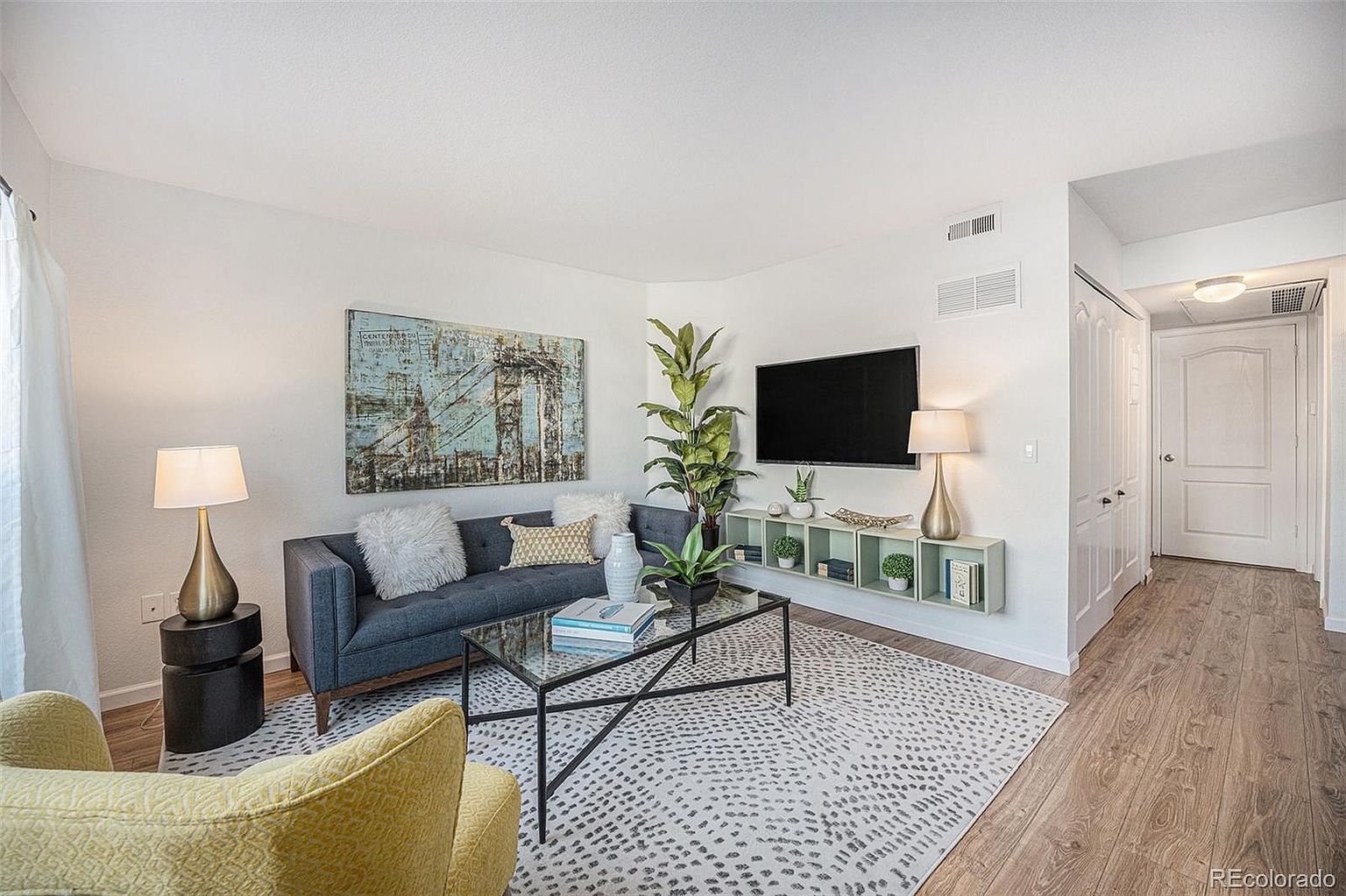 This is a well-staged living room featuring a gray sofa with decorative pillows, a glass-topped coffee table, and an accent chair. A large abstract painting hangs above the sofa, and a flat-screen TV is mounted on the wall. The room is bright and airy, with a neutral color palette and modern decor, creating a welcoming and stylish atmosphere.
