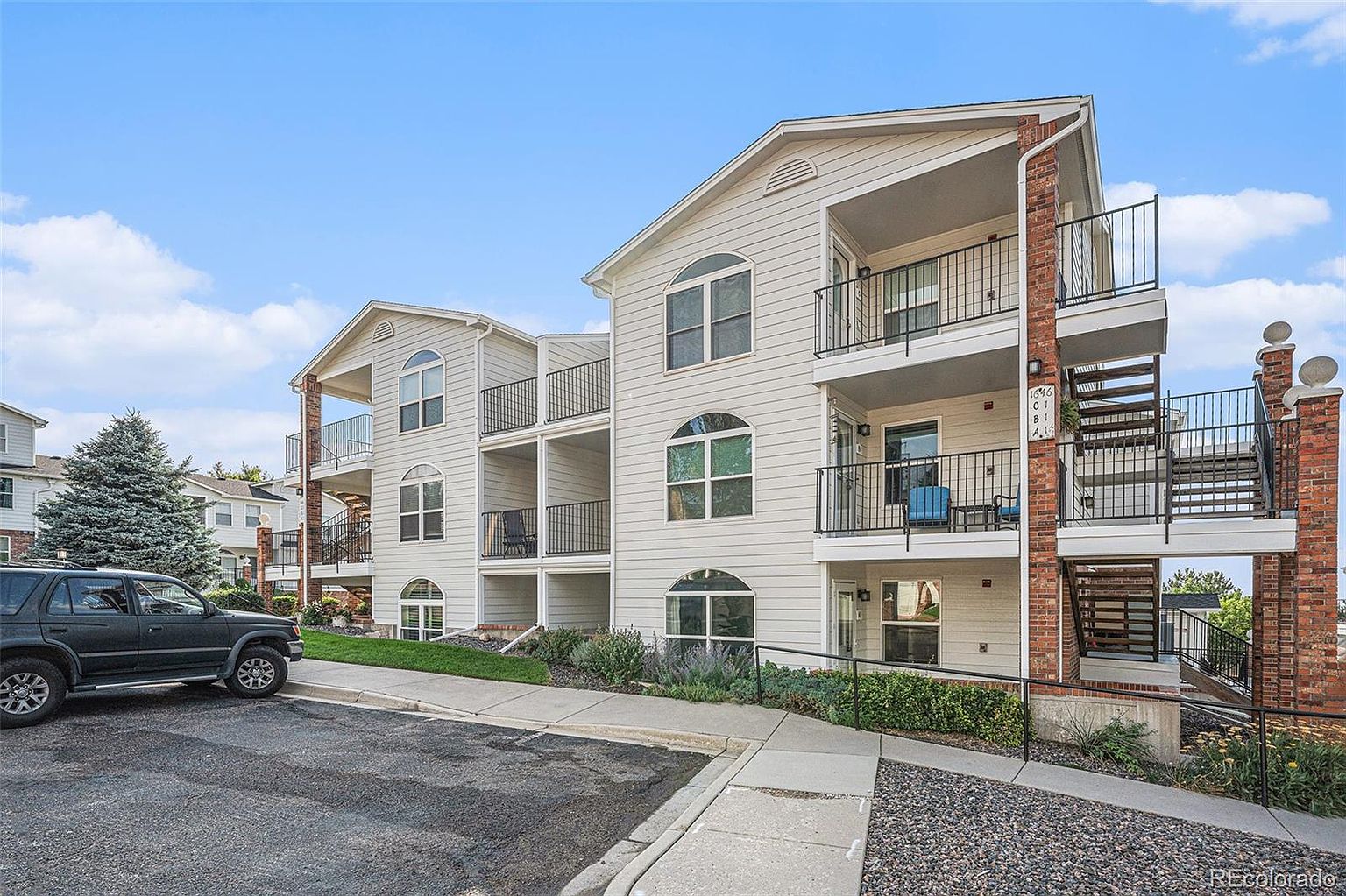 This is a front view of a multi-story apartment building with light-colored siding and brick accents. Each unit appears to have a private balcony with black metal railings. A black SUV is parked in the foreground, and landscaping adds to the curb appeal.