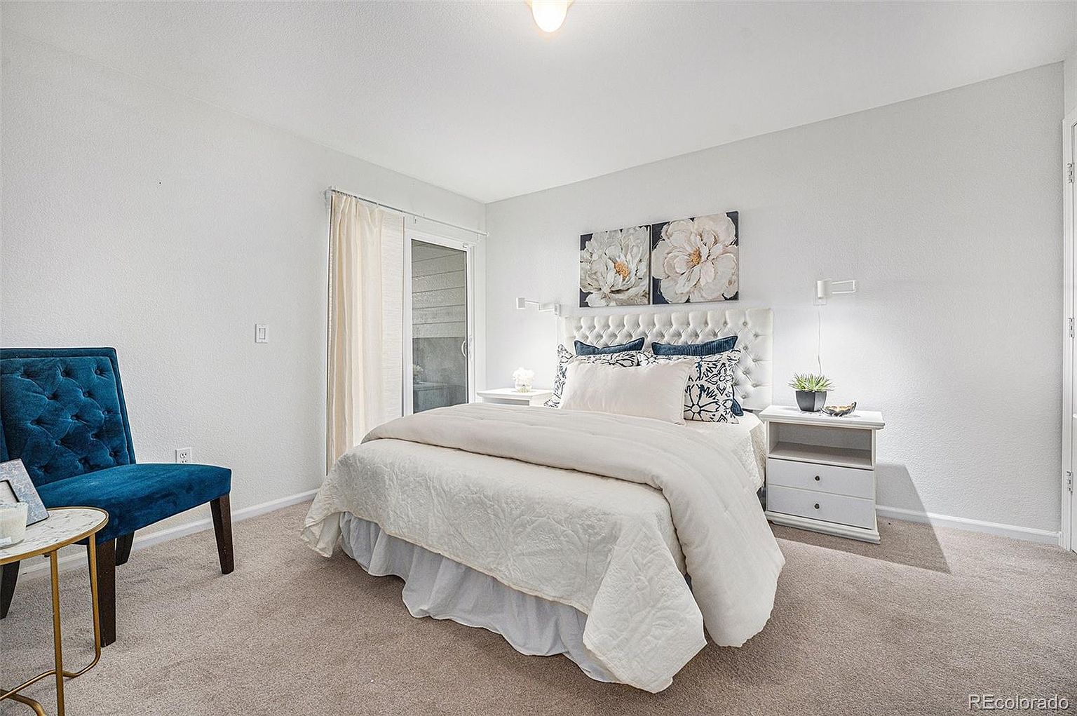 This is a bright and inviting primary bedroom featuring a white tufted headboard, crisp white bedding, and elegant floral artwork. The room is well-lit with wall sconces and natural light from a sliding glass door, complemented by a plush carpet and a stylish blue accent chair. The overall impression is clean, comfortable, and sophisticated, perfect for relaxation.