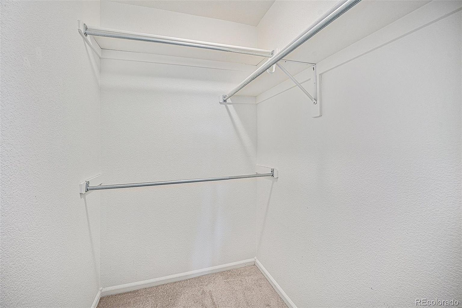 This is a well-lit, empty walk-in closet with white walls and neutral carpet. It features two metal rods for hanging clothes, one higher and one lower, providing ample storage space. The closet appears clean and ready for use, offering a blank canvas for organizing personal belongings.