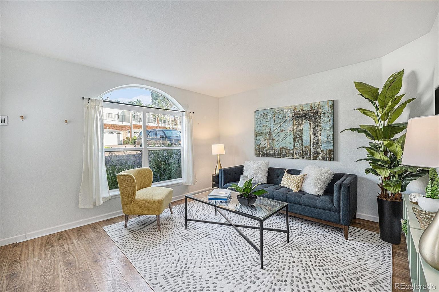 This is an interior shot of a living room featuring a blue sofa with decorative pillows, a glass-topped coffee table, and a yellow accent chair. A large arched window provides natural light, and a piece of artwork hangs above the sofa. The room is decorated in a modern style with a neutral-toned rug and hardwood flooring, creating a welcoming and stylish atmosphere.