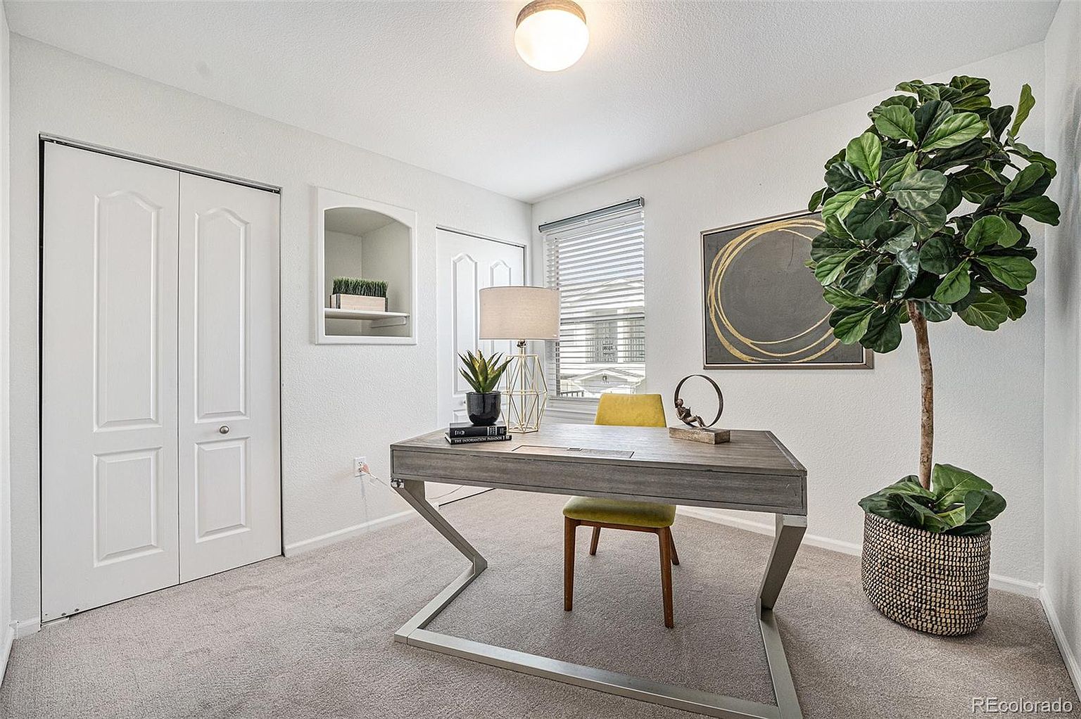 This is an interior shot of a home office. The room features a modern desk with a light wood top and silver metal legs, paired with a yellow chair. A large fiddle-leaf fig tree in a woven basket adds a touch of nature, while a piece of abstract art hangs on the wall. The room is well-lit and has a clean, contemporary feel.