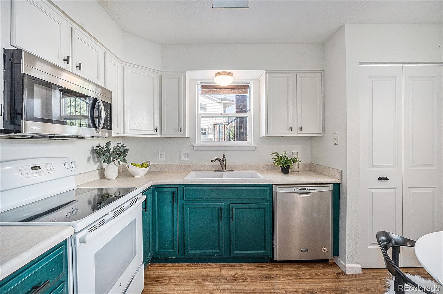 This is a well-lit kitchen featuring a combination of white and teal cabinetry, creating a bright and inviting space. Stainless steel appliances, including a microwave and dishwasher, complement the modern design. The wood-look flooring adds warmth to the room, while the window above the sink provides natural light and a view to the outside.