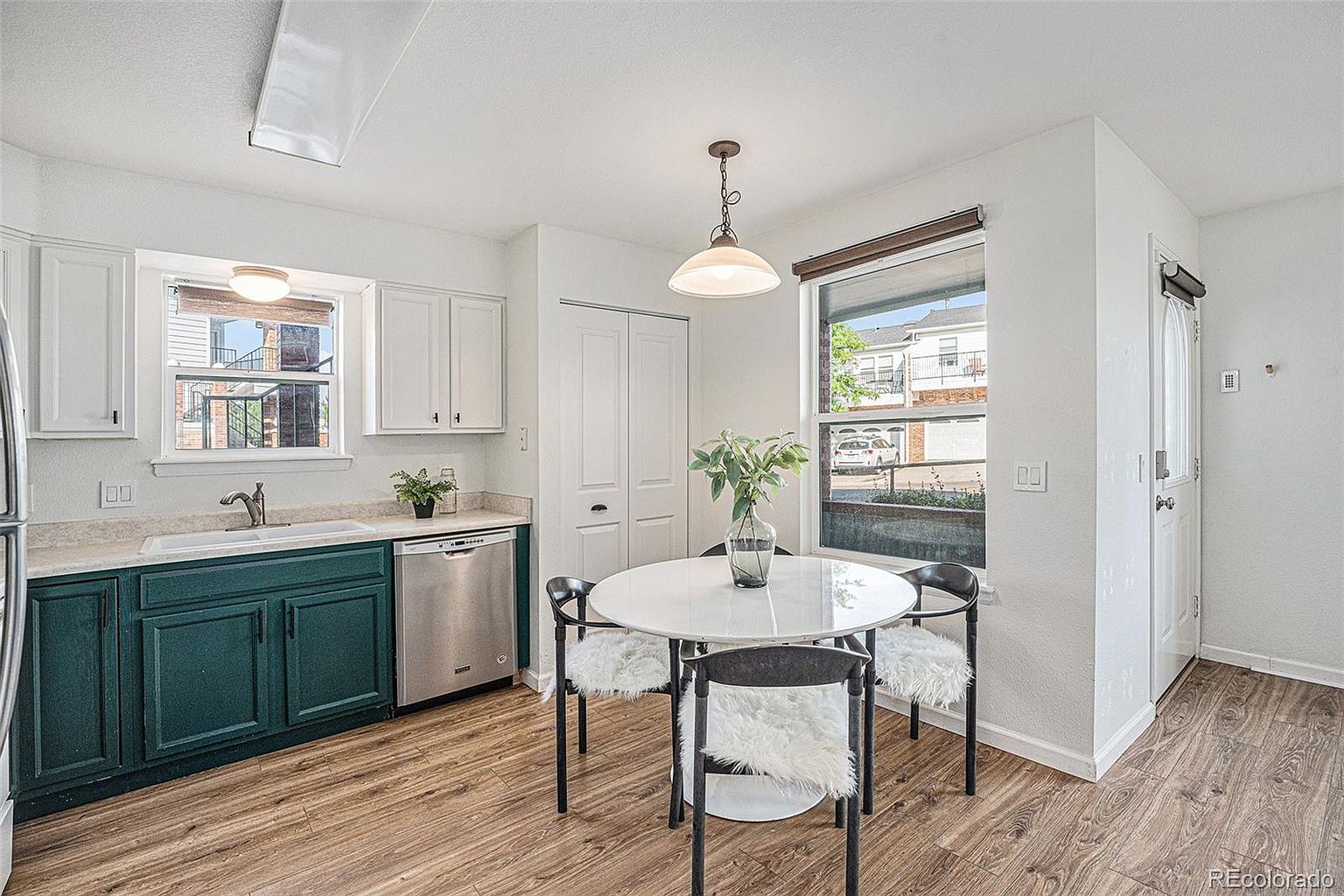This is a well-lit kitchen and dining area featuring a combination of white and dark green cabinetry, stainless steel appliances, and wood-look flooring. A round dining table with modern chairs sits near a window, creating a cozy breakfast nook. The overall impression is clean, inviting, and functional.