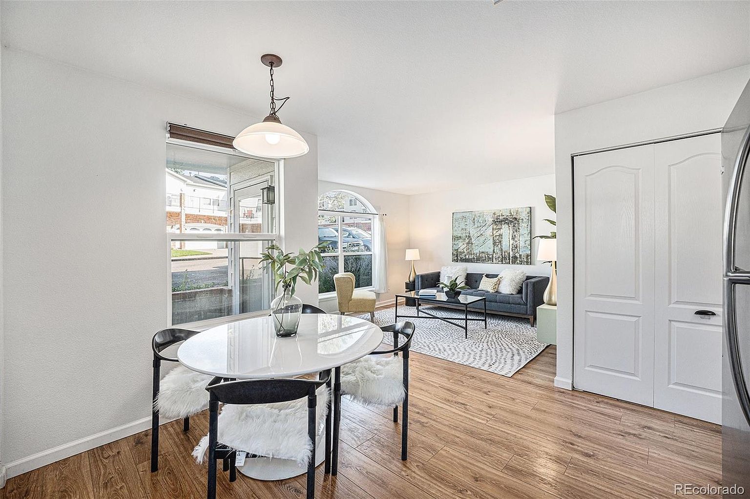 This interior shot showcases a bright and airy dining area that seamlessly transitions into a living space. The dining area features a modern round table with stylish chairs, complemented by a pendant light fixture. The living area includes a comfortable sofa, a coffee table, and a large piece of artwork, creating an inviting and cohesive living space.