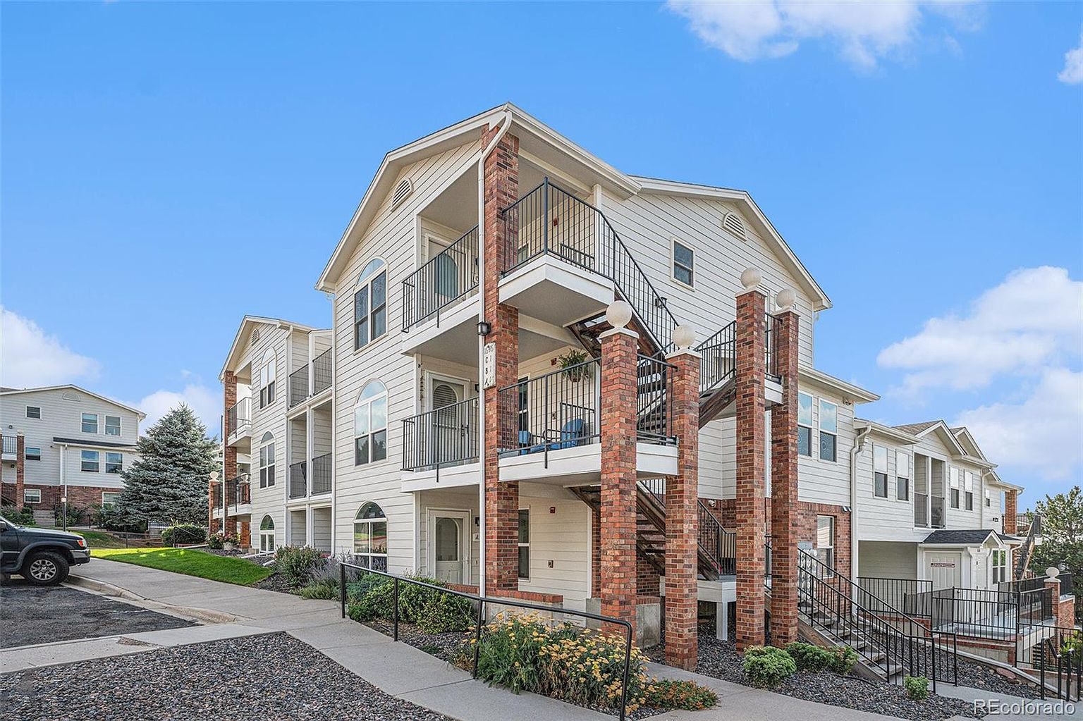 This is a front view of a multi-story apartment building with a light-colored exterior and brick accents. The building features multiple balconies with black railings and external staircases. Landscaping includes a walkway, plants, and gravel, creating a welcoming entrance.