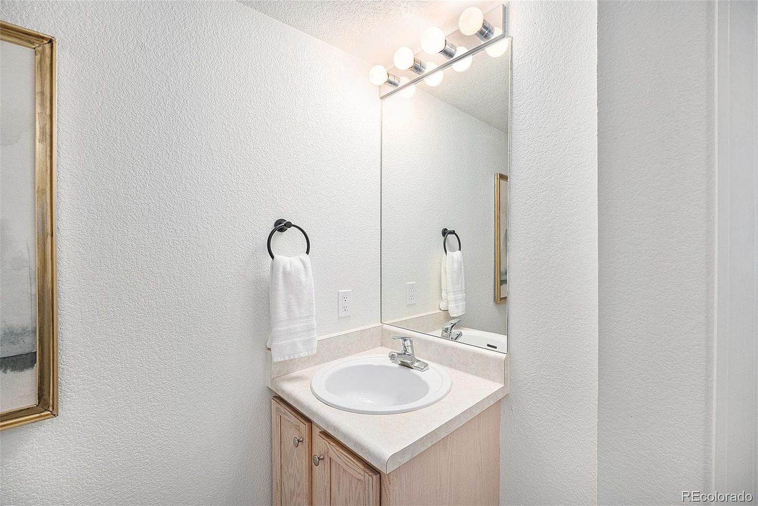 This is a well-lit guest bathroom featuring a single sink vanity with light wood cabinetry and a white countertop. A large mirror with multiple lights above it enhances the brightness of the space. A towel ring with a white towel is mounted on the wall to the left of the sink.