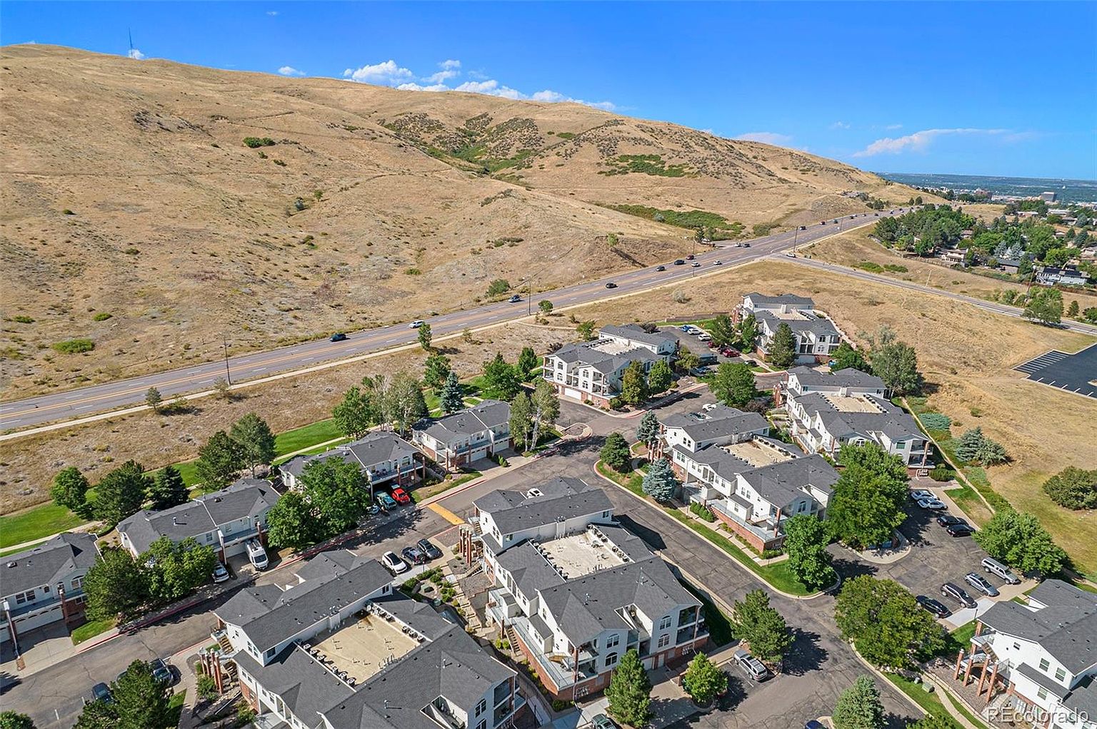 This aerial shot showcases a well-maintained residential complex nestled against a hillside. The buildings feature consistent architectural styles with gray roofs and light-colored exteriors, surrounded by mature trees and landscaping. A nearby road with light traffic adds to the suburban setting, creating a sense of community and accessibility.