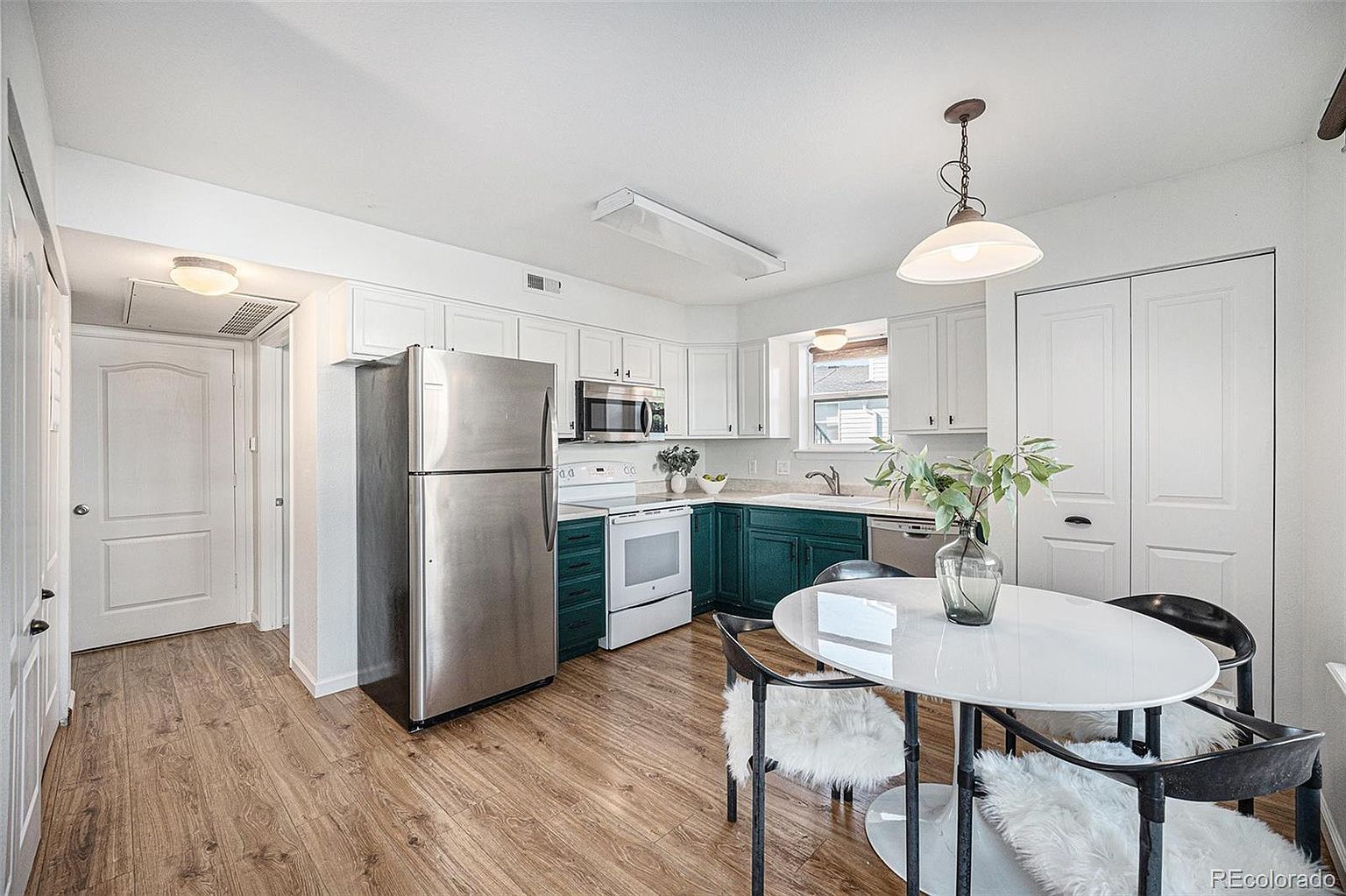This is a bright and airy kitchen featuring white upper cabinets and teal lower cabinets, complemented by stainless steel appliances. A round white dining table with modern chairs sits in the corner, illuminated by a pendant light. The wood-look flooring adds warmth to the space, creating an inviting atmosphere.