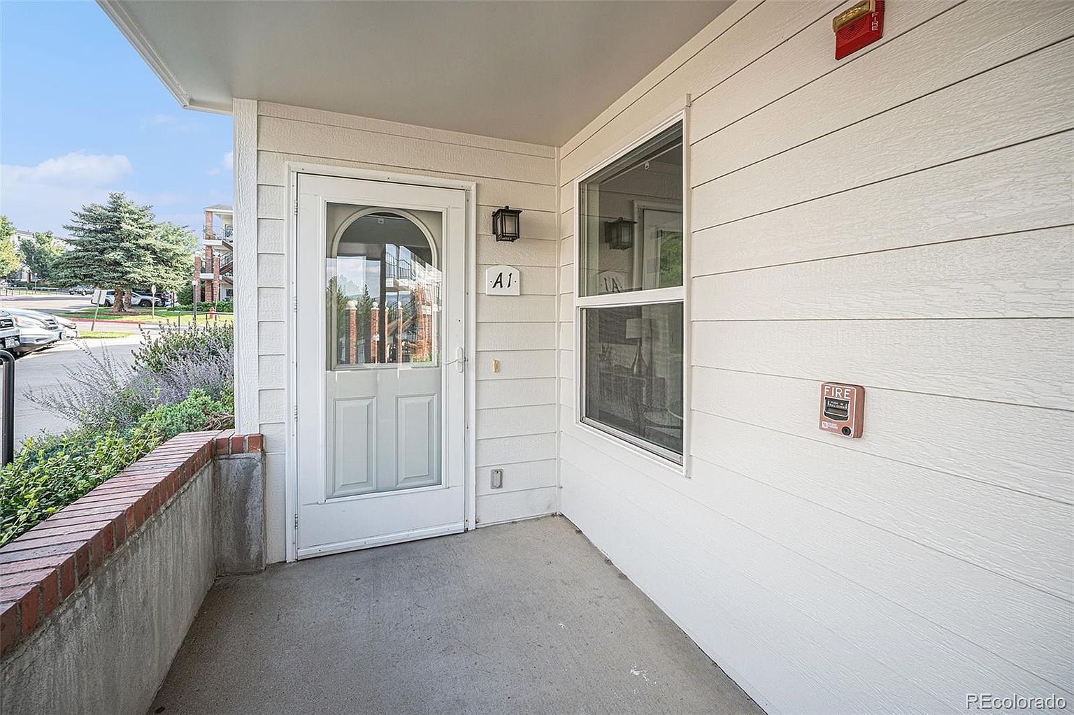 This is an exterior shot of a home's entryway, featuring a white door with a glass panel and the unit number "A1" displayed above. The entrance area has a concrete floor and a low brick wall to the side, with a window on the right side of the door. The siding is light-colored, and there's a fire alarm pull station visible on the wall.