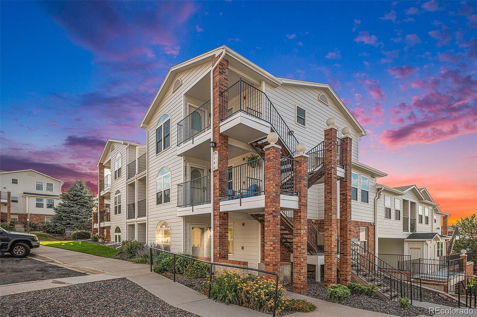 This is a front view of a multi-story apartment building with a light-colored exterior and brick accents. Each unit appears to have a private balcony accessible by an external staircase. The building is well-maintained with landscaping and a paved walkway, suggesting a comfortable and inviting living environment.