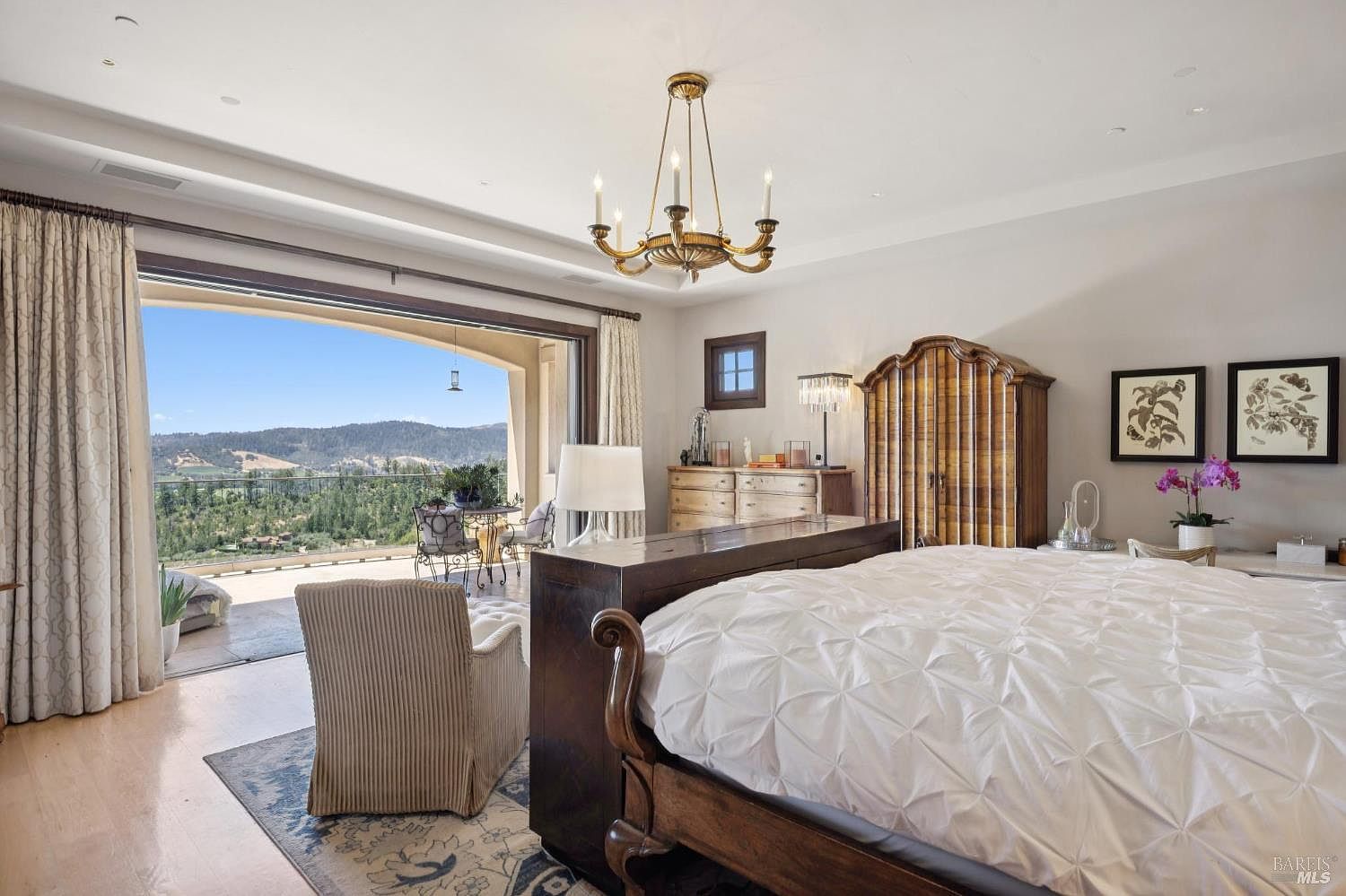 This elegant primary bedroom features a large wooden bed frame with a tufted white comforter and a cozy armchair positioned on a patterned area rug. The room opens onto a spacious balcony through expansive glass doors, offering panoramic views of the surrounding hills and landscape. A classic chandelier hangs from the ceiling, while a tall wooden armoire and framed botanical art add a touch of traditional sophistication to the light-filled space.