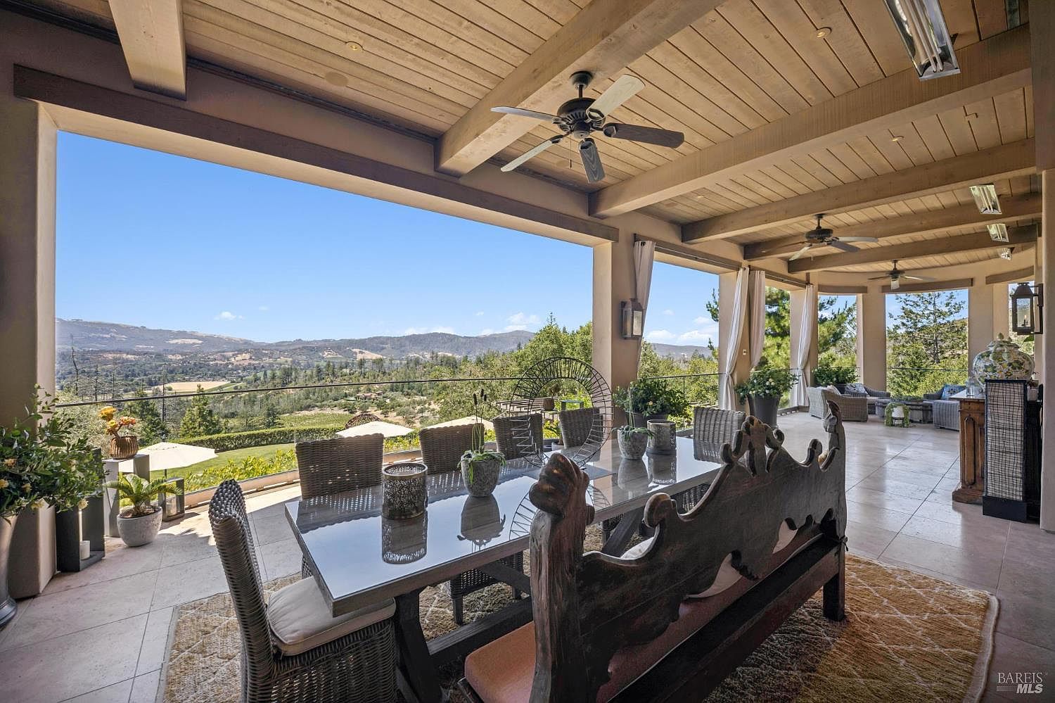 This expansive covered patio offers a sophisticated outdoor dining and lounge area with panoramic views of rolling hills and vineyards. The space features a large dining table with a unique carved wooden bench, tile flooring, and multiple ceiling fans, creating an ideal setting for al fresco entertaining. The open-air design seamlessly connects the indoor-outdoor living experience, framed by sturdy pillars and a natural wood-paneled ceiling.