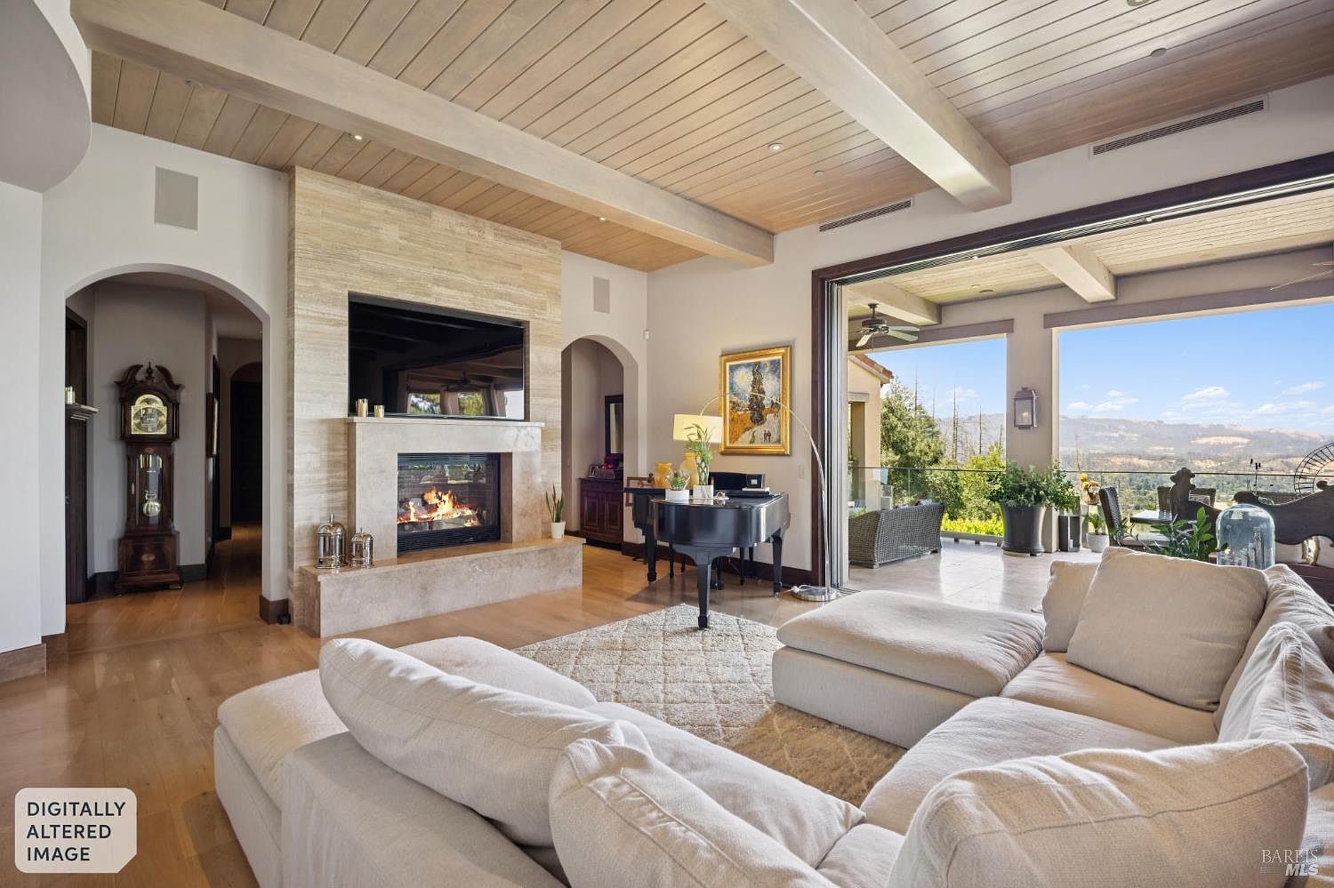 This spacious living room features a light-toned wood-paneled ceiling with exposed beams and a large stone-clad chimney breast housing a fireplace and television. A grand piano sits near the transition to an expansive outdoor patio, which is accessed through wide sliding glass doors that frame a scenic mountain view. The room is furnished with a large, plush sectional sofa and a grandfather clock, creating an elegant and inviting atmosphere.