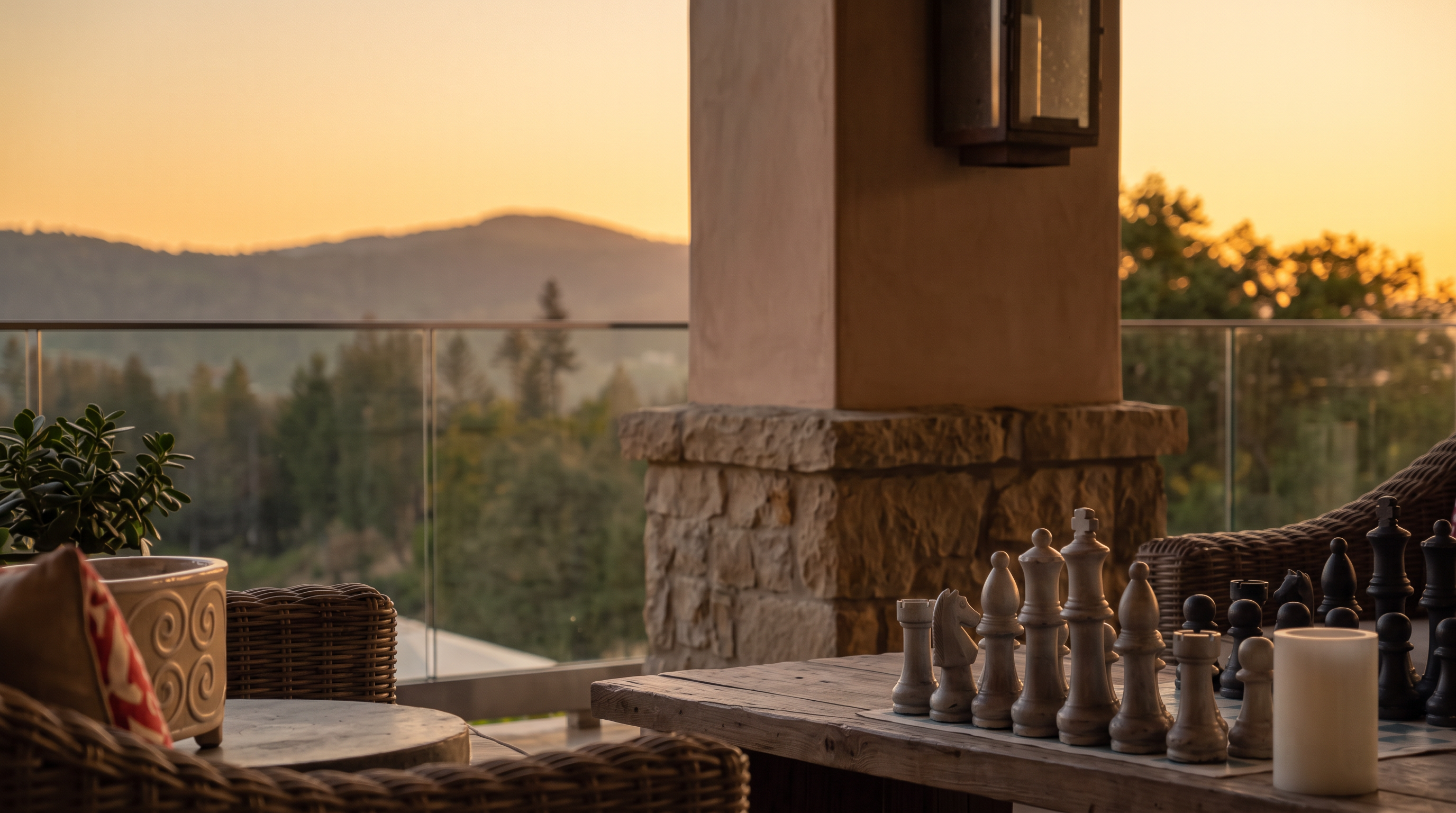 This expansive covered patio offers a luxurious outdoor living space with panoramic views of the surrounding hills and forest. The area is furnished with comfortable wicker seating featuring red patterned cushions, a rustic wooden coffee table with a chess set, and a formal dining arrangement further down the deck. The space is defined by a high, wood-paneled ceiling with multiple fans and a sleek glass railing that maintains an unobstructed connection to the natural landscape.