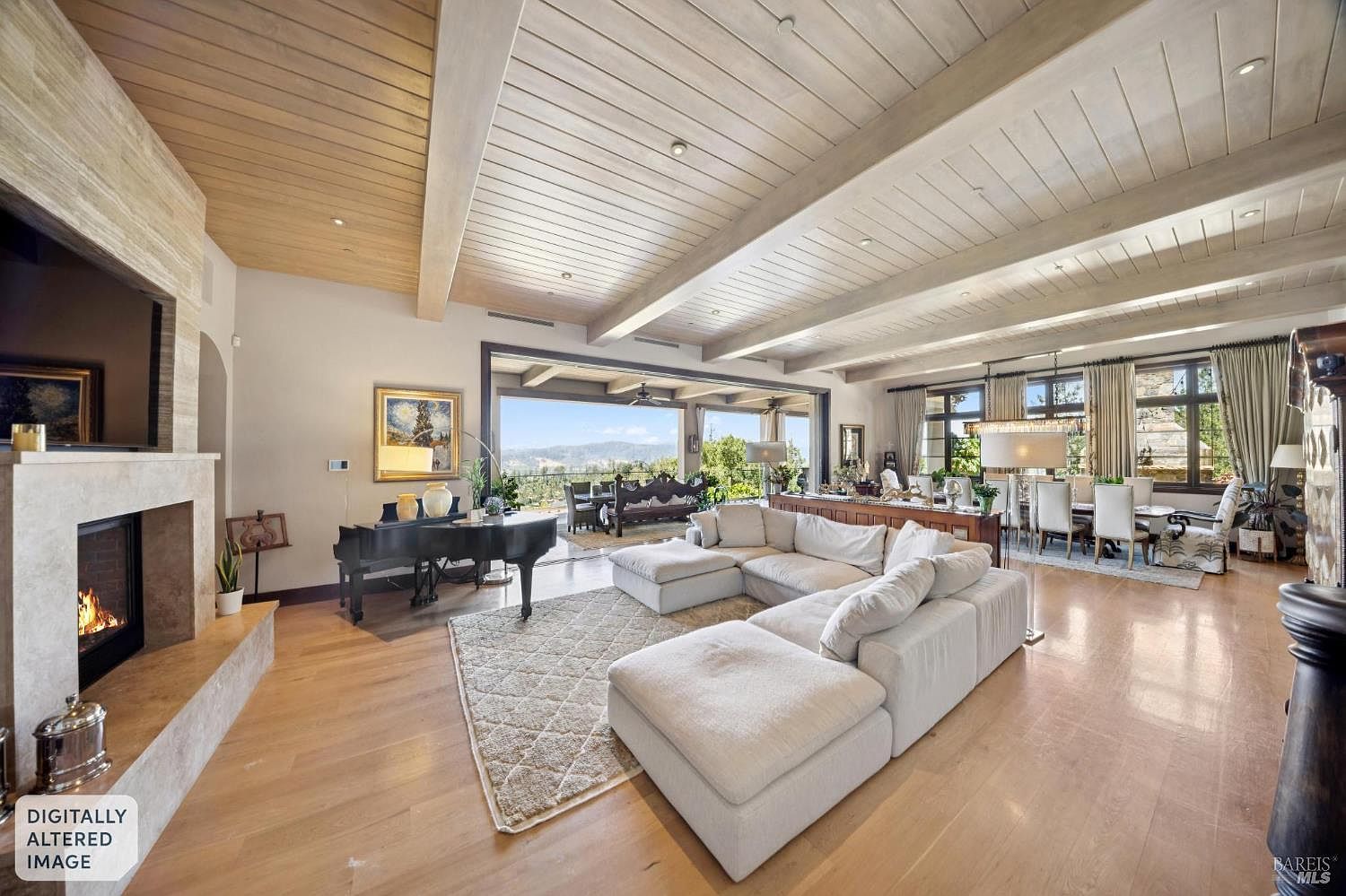 This expansive open-concept living area features light wood flooring and a vaulted ceiling with exposed beams. A large, plush sectional sofa faces a stone-clad chimney breast with a built-in fireplace, while a grand piano sits nearby. The space flows seamlessly toward a wide opening that reveals a scenic outdoor view, creating a bright and airy atmosphere suitable for luxury entertaining.