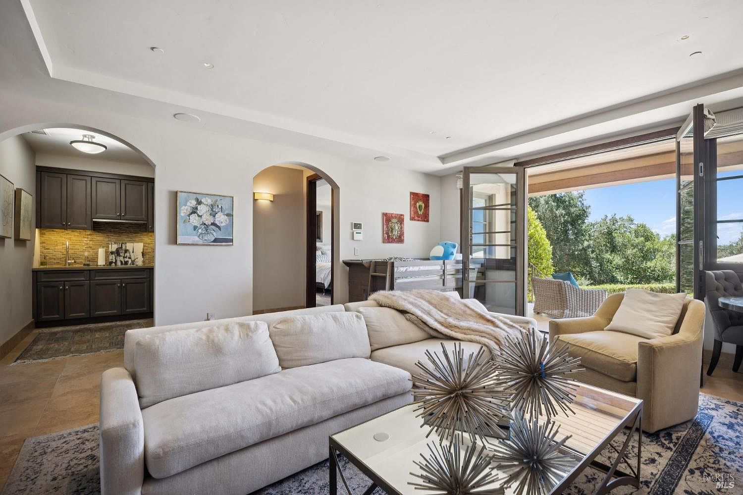 This bright and inviting living room features a large, plush sectional sofa and a contemporary glass-topped coffee table adorned with metallic starburst sculptures. The space flows seamlessly into a wet bar area with dark cabinetry and an arched entryway, while large glass doors open to a sun-drenched patio with outdoor seating. The neutral color palette and soft textures create a sophisticated, comfortable atmosphere perfect for relaxation or entertaining.
