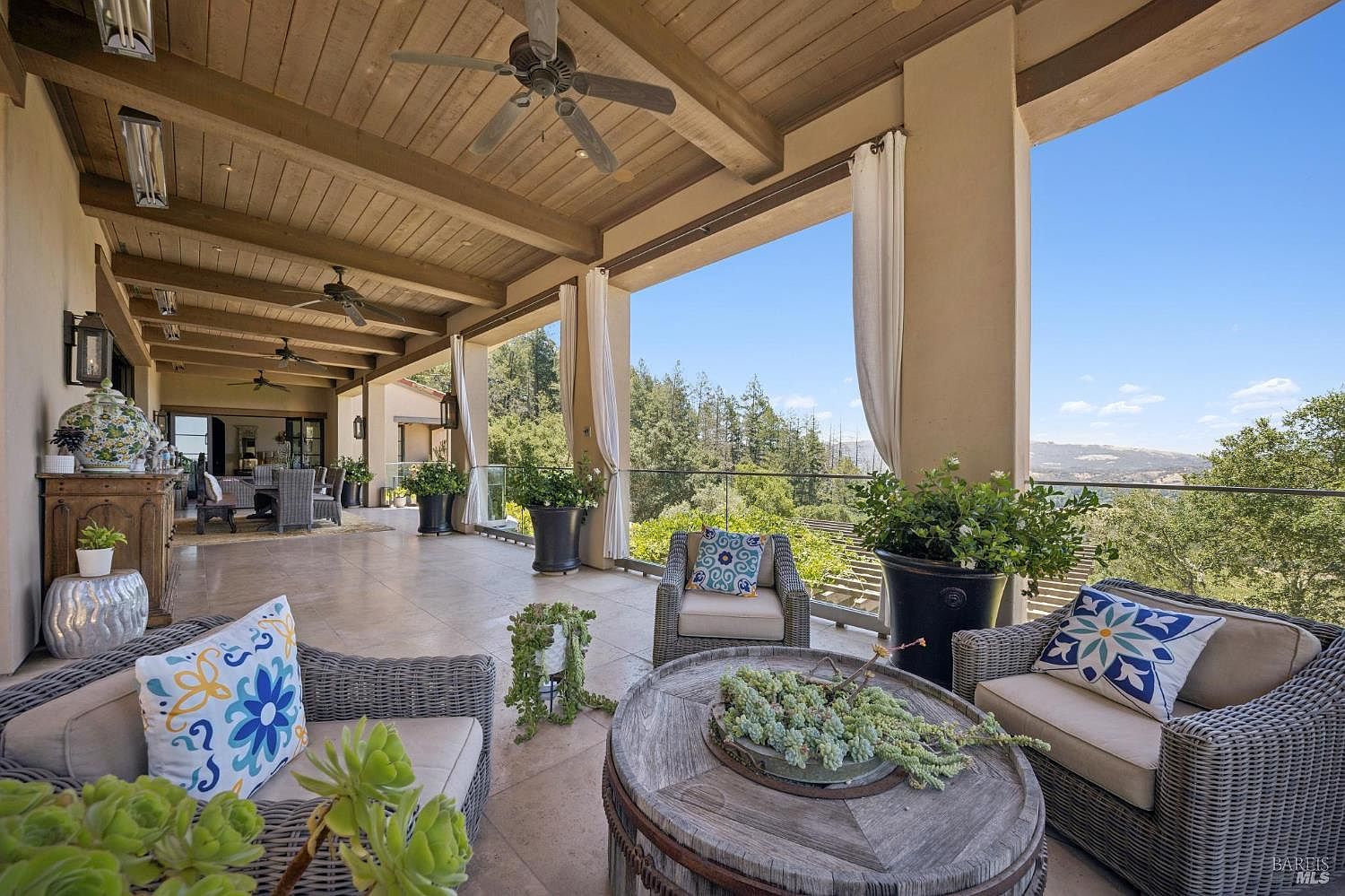 This expansive covered patio offers a luxurious outdoor living space with a panoramic view of the surrounding hills and trees. The area is furnished with comfortable wicker seating, a rustic circular wooden coffee table, and several potted plants, creating a serene and inviting atmosphere. The space features a wood-paneled ceiling with multiple ceiling fans and recessed lighting, providing a seamless transition between indoor comfort and the natural landscape.