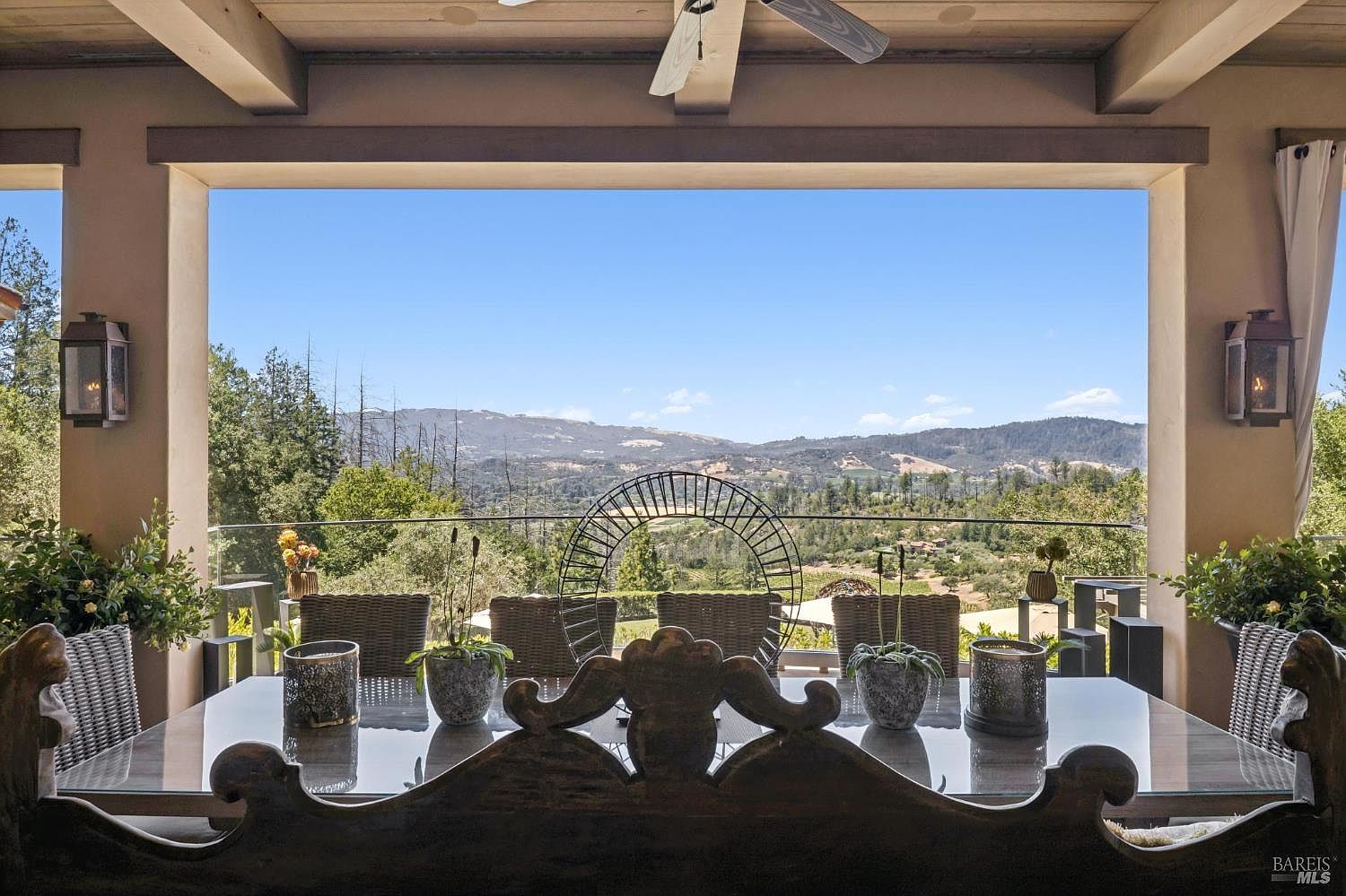 This covered outdoor dining area offers a stunning, elevated view of rolling hills and lush landscape. The space features a large glass-topped table with woven chairs, framed by a sleek glass railing that ensures an unobstructed vista. The architectural design includes a wooden ceiling with a central fan and wall-mounted lanterns, creating a sophisticated and inviting atmosphere for al fresco dining.