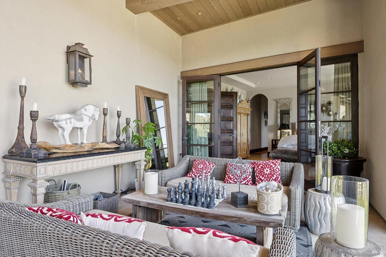 This covered outdoor living space features a sophisticated, rustic-chic aesthetic with wicker seating and a weathered wood coffee table topped with a decorative chess set. A console table against the wall displays a white horse sculpture and candle holders, while large glass doors provide a seamless transition into the adjacent interior bedroom. The space is defined by a wood-paneled ceiling and neutral walls, creating a serene and inviting atmosphere for relaxation.