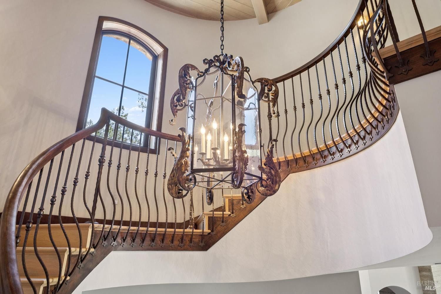 This image captures a grand, curved staircase featuring ornate dark metal railings and light wood treads. A large, intricate metal lantern-style chandelier hangs centrally, serving as a dramatic focal point within the stairwell. To the left, a tall, arched window allows natural light to flood the space, highlighting the elegant architectural curves and the neutral-toned walls.