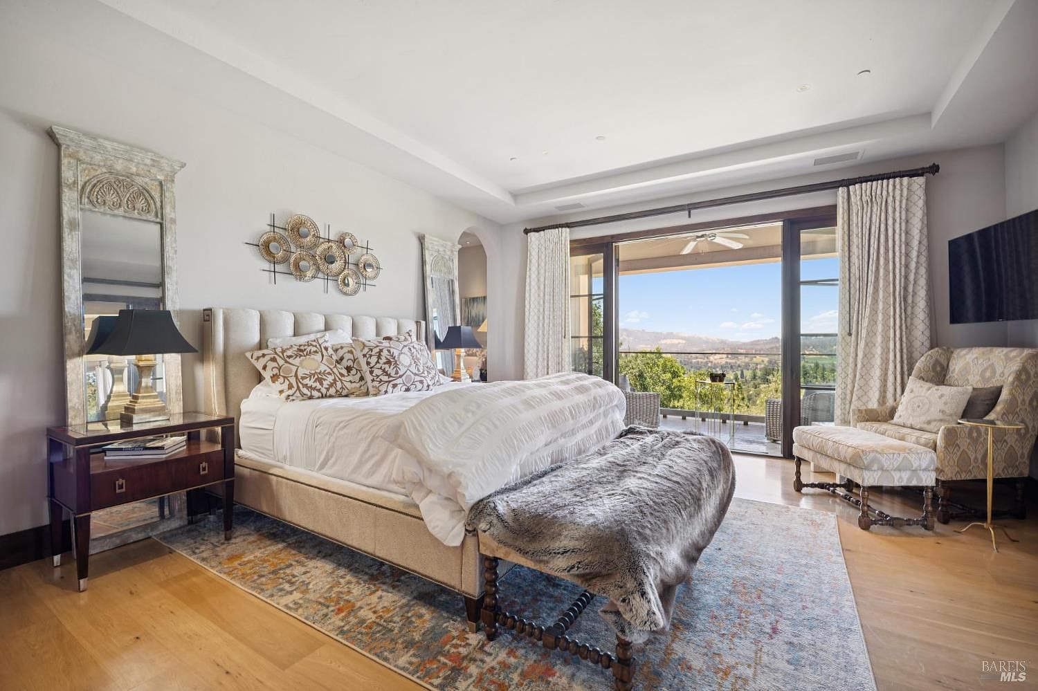 This elegant primary bedroom features a plush upholstered bed topped with patterned pillows and a faux-fur throw, centered on a decorative area rug over light wood flooring. Large glass sliding doors open to a private balcony, offering expansive views of the surrounding landscape and hills. The room is accented by tall mirrors, a patterned armchair, and a unique metallic wall art piece, creating a sophisticated and serene atmosphere.