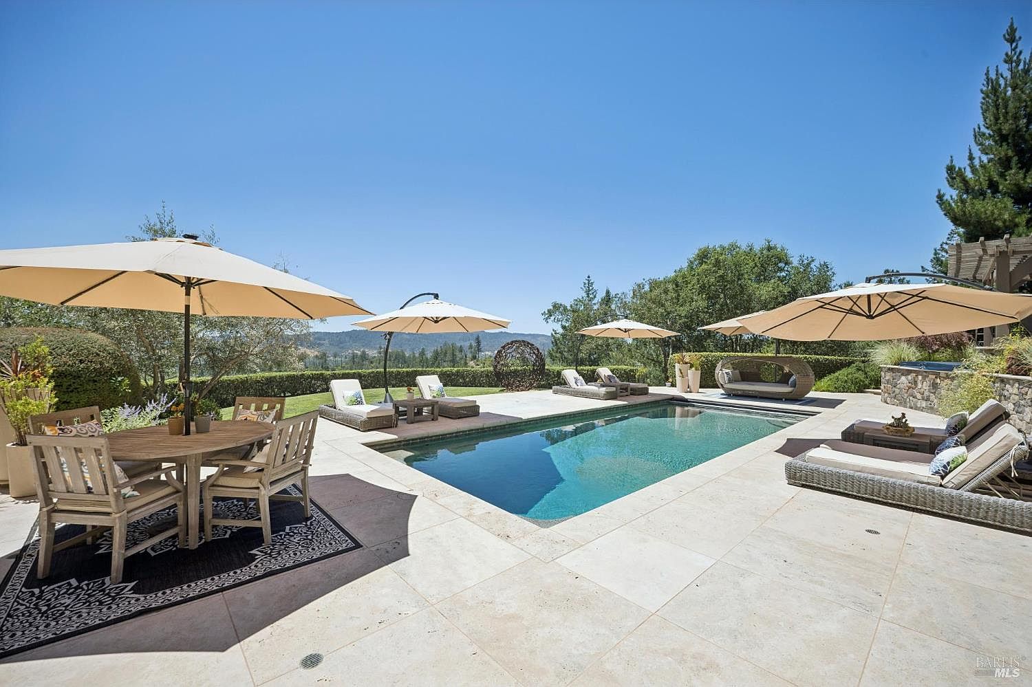 This expansive outdoor living space features a rectangular swimming pool as the centerpiece, surrounded by light-toned stone decking. The area is furnished with multiple lounge chairs, a dining set under a large umbrella, and a unique circular daybed, all set against a backdrop of rolling hills and mature trees. The scene captures a serene, resort-style atmosphere perfect for entertaining and relaxation.