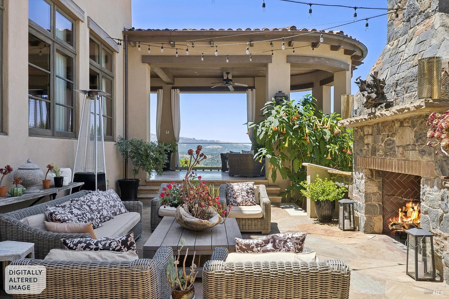 This inviting outdoor living space features a comfortable seating area arranged around a central coffee table, set against a backdrop of a stone-clad chimney breast with a roaring fire. The patio is partially covered by a pergola structure adorned with string lights, leading to an elevated deck area that offers expansive views of the surrounding landscape. The space is decorated with potted plants and plush, patterned cushions, creating a warm and sophisticated atmosphere for relaxation or entertaining.