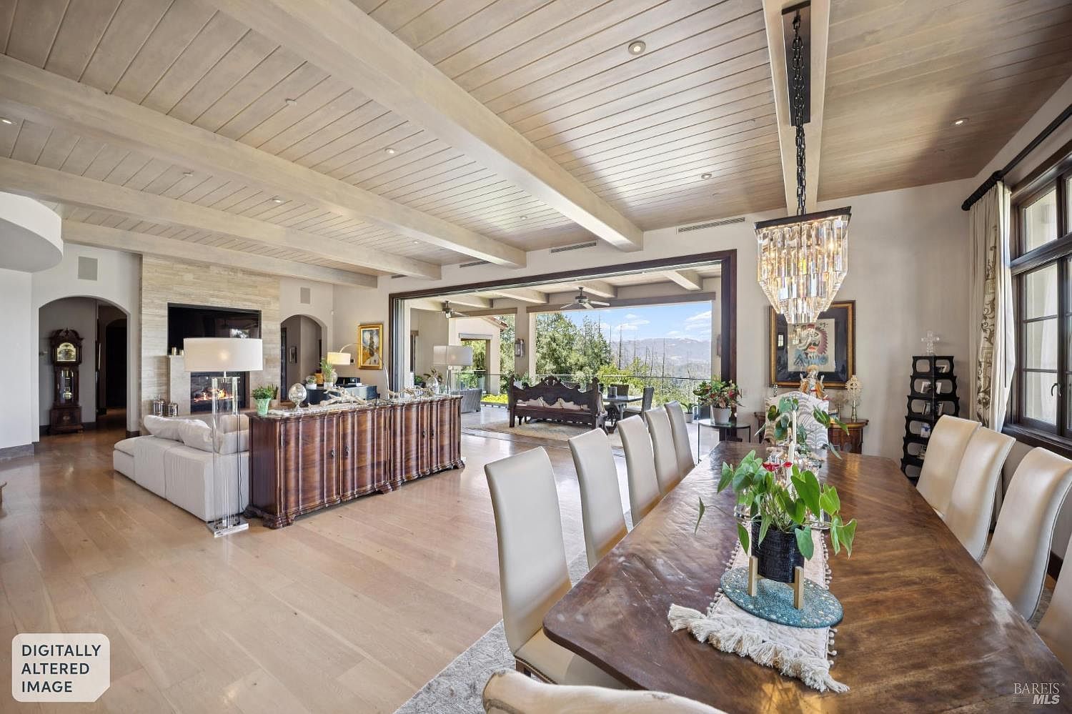 This expansive open-concept living and dining area features light wood flooring and a vaulted ceiling with exposed beams. A large dining table sits in the foreground under a crystal chandelier, while a comfortable living area with a stone-clad fireplace and a long wooden console table occupies the background. The space flows seamlessly through a wide opening to a covered outdoor patio, offering a panoramic view of the surrounding landscape.