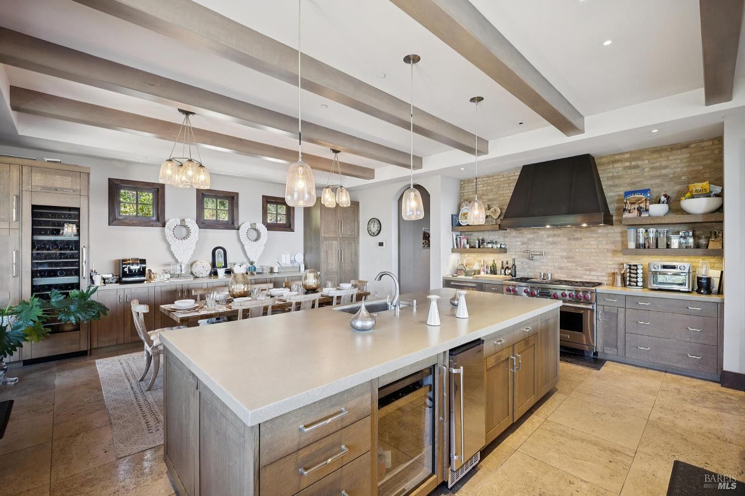 This spacious, open-concept kitchen features a large central island with a stone countertop, integrated appliances, and a sink, serving as the focal point of the room. The space is defined by exposed wooden ceiling beams, light-toned cabinetry, and a textured brick wall behind the professional-style range and hood. A dining area is seamlessly integrated into the space, creating a warm and inviting atmosphere suitable for both cooking and entertaining.