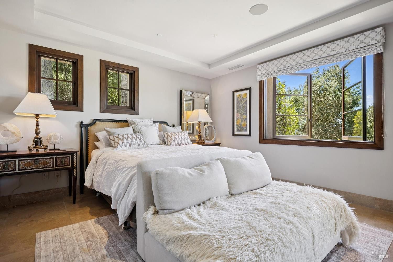 This elegant primary bedroom features a neutral color palette with a plush, upholstered bed frame and a textured, faux-fur bench at the foot. The room is accented by dark wood-framed windows, a decorative side table with a classic lamp, and a large mirror reflecting the space. A wide window with a patterned roman shade provides a view of the surrounding trees, creating a serene and inviting atmosphere.