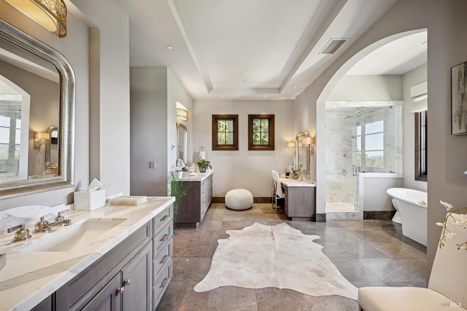 This spacious primary bathroom features dual vanities with stone countertops, a dedicated makeup vanity area, and a large walk-in glass shower. A freestanding soaking tub is positioned near a window, while a light-colored animal hide rug rests on the center of the tiled floor. The room is finished with neutral tones, elegant lighting fixtures, and a bright, airy atmosphere.