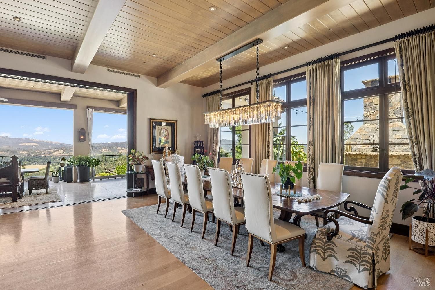 This elegant dining room features a long wooden table surrounded by upholstered chairs, set beneath a prominent rectangular crystal chandelier. The space is defined by a light-toned wood plank ceiling and large glass doors that open to a covered patio, creating a seamless transition to the outdoor landscape. A patterned area rug anchors the dining area, while the room's neutral color palette and natural light contribute to a sophisticated and airy atmosphere.