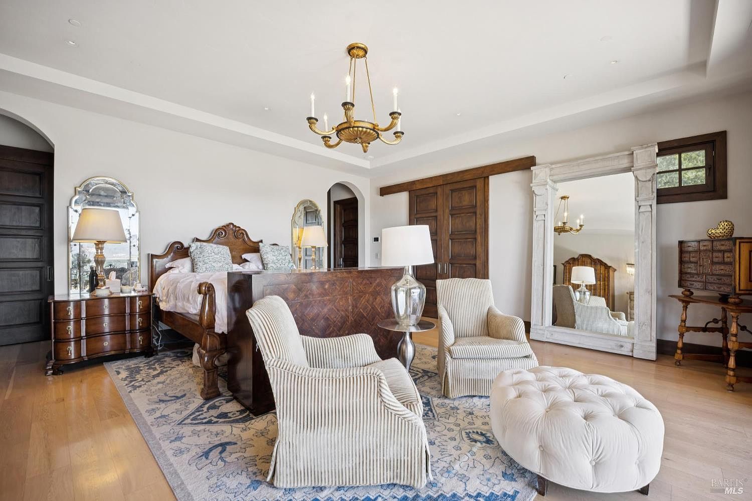 This spacious primary bedroom features a classic, elegant design with a large wooden bed frame positioned in the center of the room, backed by a dark wood console. The space is furnished with two striped armchairs, a tufted ottoman, and a large floor mirror, all set atop a patterned area rug on light wood flooring. A gold chandelier hangs from the ceiling, and the room is accented by arched doorways and dark wood sliding doors, creating a sophisticated and comfortable atmosphere.