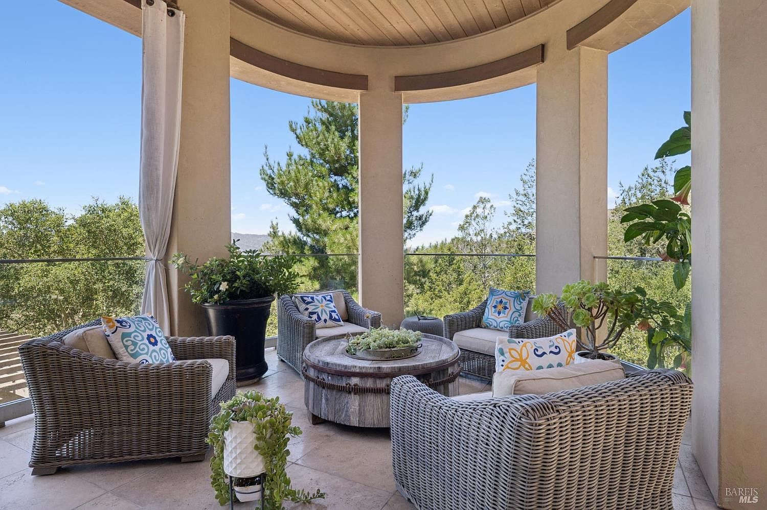 This covered balcony offers a serene outdoor living space with a curved architectural design and expansive views of the surrounding trees and hills. The area is furnished with comfortable wicker-style seating arranged around a rustic, circular wooden coffee table, creating an inviting atmosphere for relaxation. The space features neutral-toned tile flooring and tall, open arches that frame the natural landscape, emphasizing a seamless connection between the home and the outdoors.