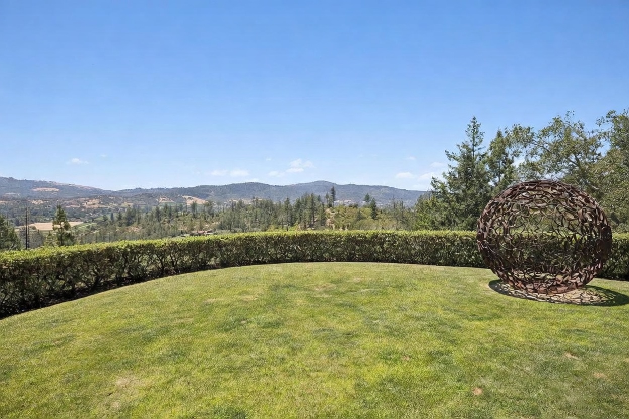 This expansive, manicured lawn offers a serene outdoor space framed by a neatly trimmed hedge that separates the yard from a breathtaking panoramic view of rolling hills and distant mountains. A large, spherical metal sculpture serves as a striking focal point on the right side of the frame, adding an artistic element to the natural landscape. The perspective is wide and elevated, capturing the vastness of the horizon under a clear, bright blue sky.