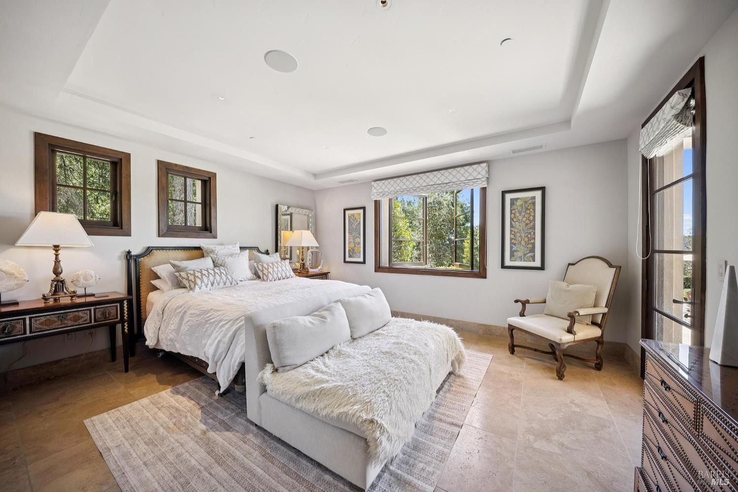 This spacious primary bedroom features a neutral color palette, stone flooring, and a large bed centered against a wall with two symmetrical windows. A plush, light-colored bench sits at the foot of the bed, while a classic armchair is positioned near a large window that offers natural light. The room is accented with framed artwork and a dark wood side table, creating an elegant and serene atmosphere.
