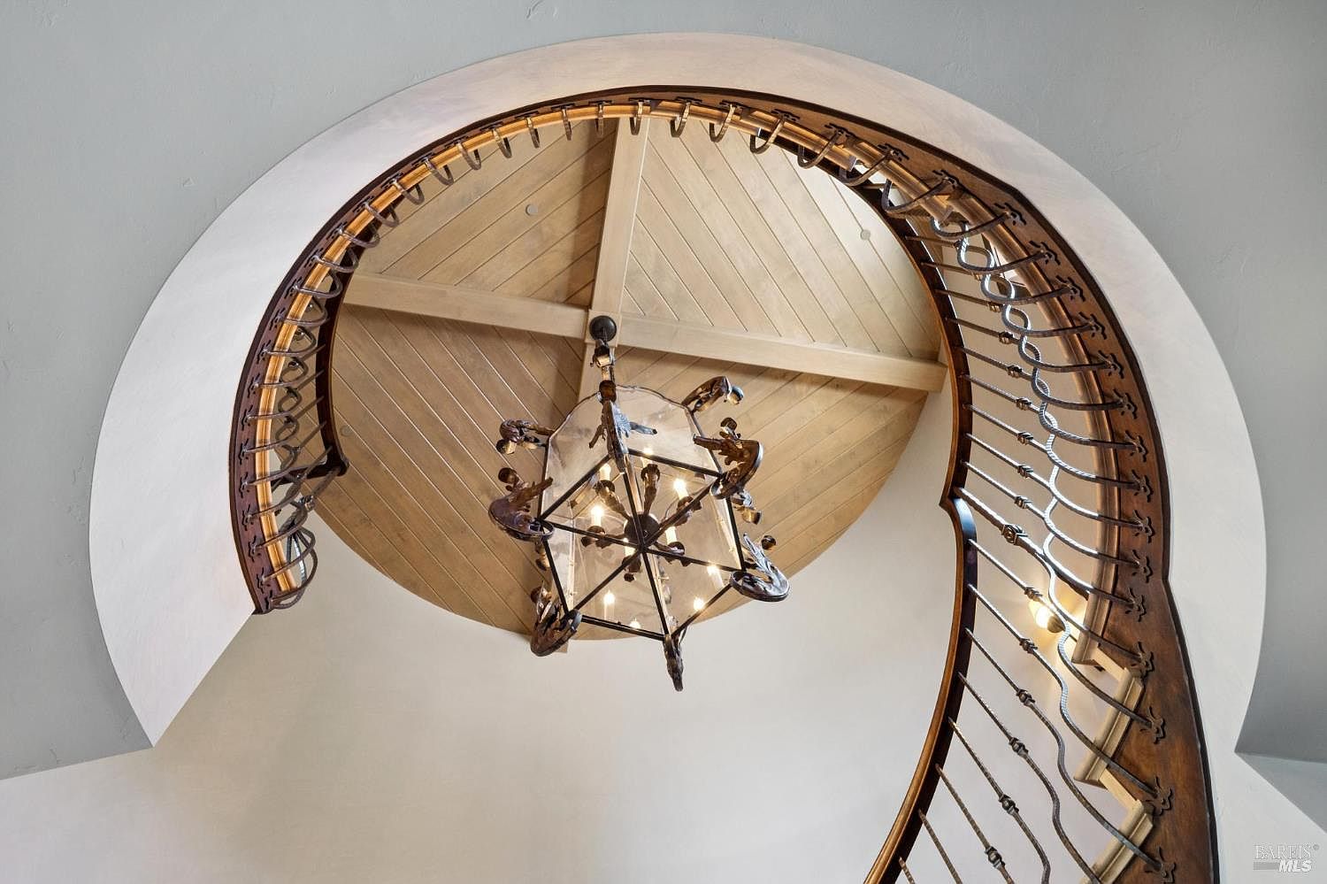 This low-angle, upward-looking perspective captures a grand, curved staircase featuring intricate dark metal railings and a warm wood handrail. Above, a large, ornate lantern-style chandelier hangs from a circular, light-wood paneled ceiling detail that adds architectural depth. The clean, neutral-toned walls provide a bright backdrop, emphasizing the elegant, sweeping lines of the staircase design.