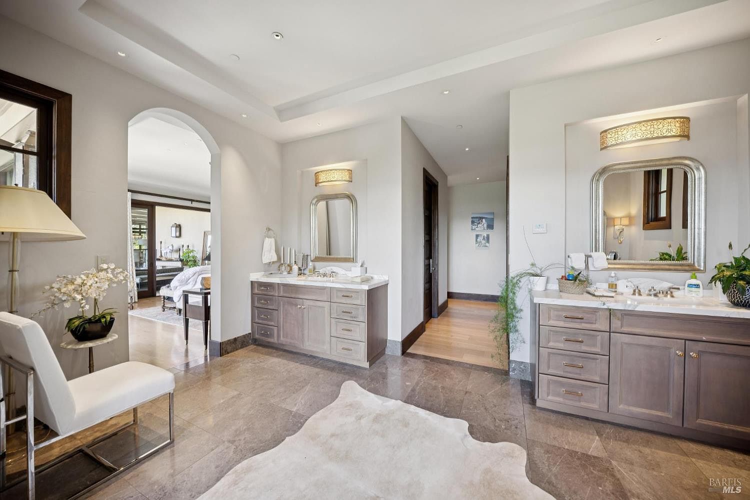 This spacious primary bathroom features dual vanities with stone countertops and light-toned cabinetry, complemented by large mirrors and decorative lighting fixtures. The room is finished with polished stone flooring and a central animal-hide rug, creating a luxurious and serene atmosphere. An arched doorway leads into an adjacent bedroom, providing a seamless flow between the living spaces.