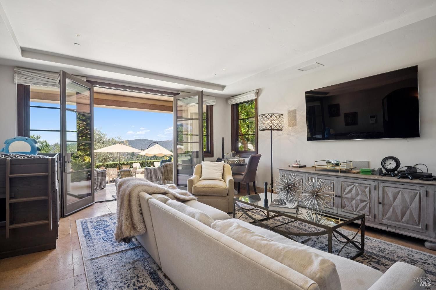 This bright and airy living room features a neutral color palette, a large sectional sofa, and a glass-topped coffee table adorned with decorative metal spheres. A large wall-mounted television sits above a rustic-style wooden media console, while tall glass doors open to a scenic outdoor patio with mountain views. The space is accented by a patterned area rug and a modern floor lamp, creating a comfortable and inviting atmosphere.
