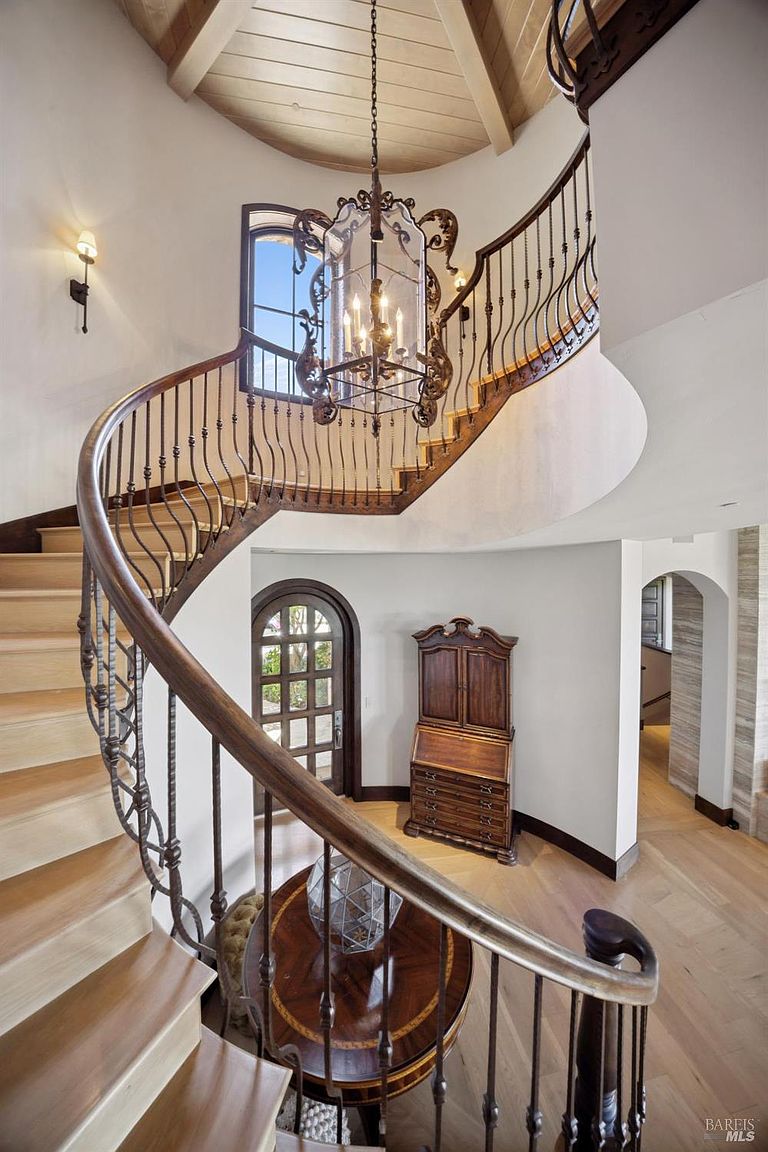 This grand foyer features a sweeping curved staircase with intricate metal railings and light wood treads. A large, ornate multi-tiered chandelier hangs from the vaulted ceiling, casting light over the entryway below where a dark wood secretary desk and a round table are positioned. The space is characterized by its elegant architectural curves, neutral walls, and a warm, inviting atmosphere.