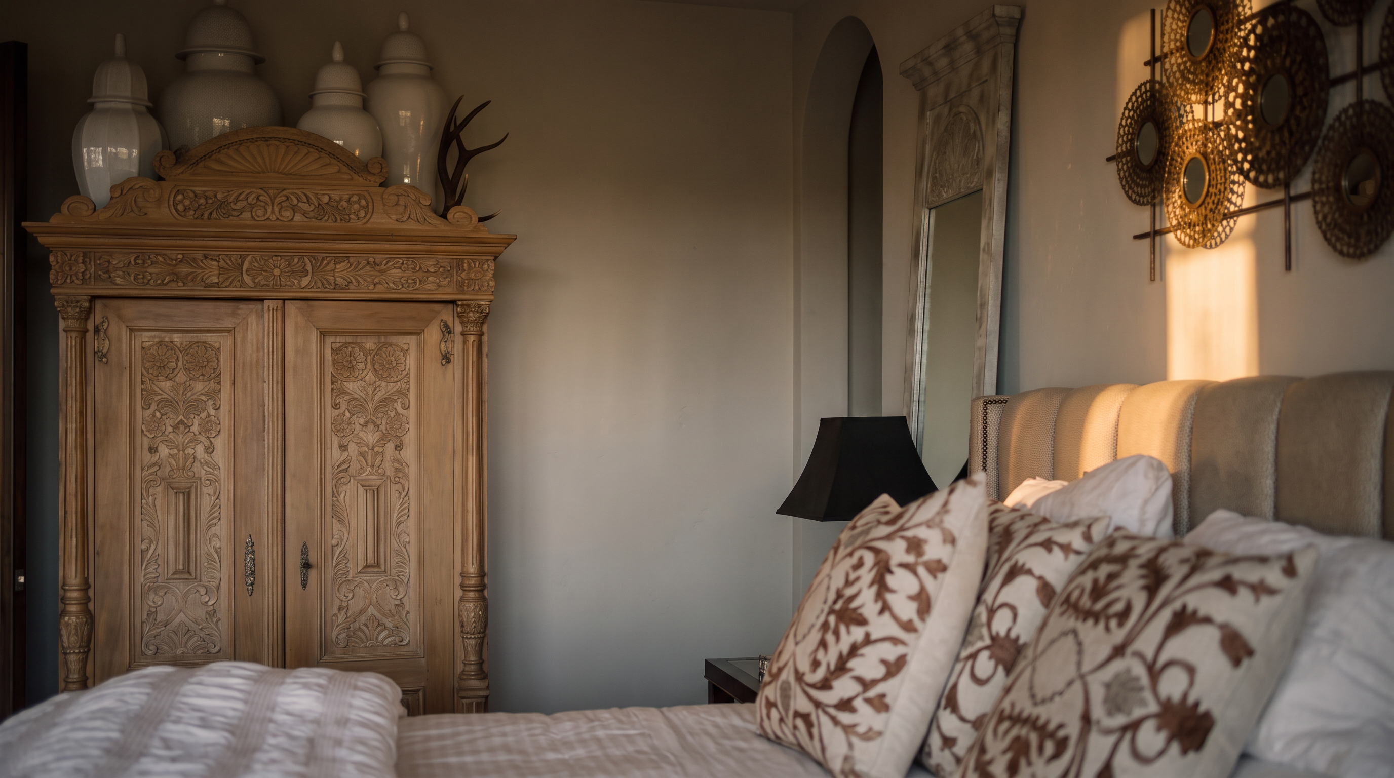 This spacious primary bedroom features a neutral color palette with a large upholstered bed as the focal point, accented by patterned throw pillows and a plush white duvet. A decorative wooden armoire stands to the left, while a tall, ornate mirror sits behind the bedside table, adding a touch of elegance to the room. The space is grounded by a colorful, patterned area rug over light wood flooring, creating a warm and inviting atmosphere.