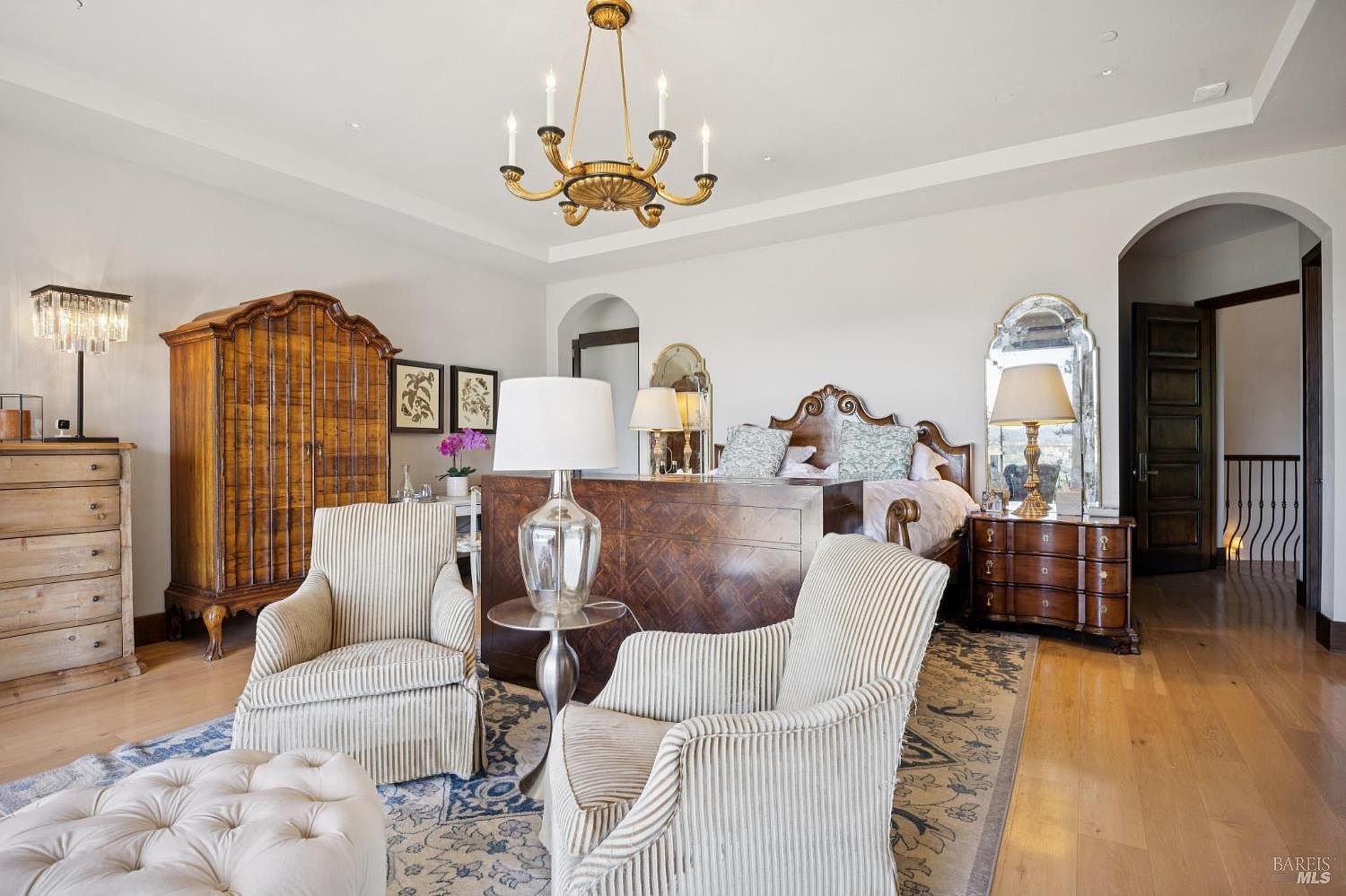 This spacious primary bedroom features a traditional aesthetic with a large wooden bed frame, a matching armoire, and a pair of striped armchairs arranged on a patterned area rug. The room is illuminated by a classic chandelier and bedside lamps, while light wood flooring and neutral walls create a bright, airy atmosphere. A tufted ottoman sits in the foreground, and an arched doorway leads to an adjacent space, providing a sense of depth and architectural interest.