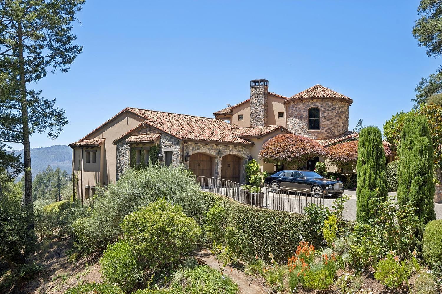 This Mediterranean-style estate features a multi-level design with a stone-clad exterior, a terracotta tile roof, and a prominent circular tower element. A paved driveway leads to a two-car garage, while a black metal railing borders the elevated terrace overlooking a lush, landscaped hillside. The scene is captured from a slightly elevated perspective, highlighting the home's architectural character and its integration into the natural, sun-drenched landscape.