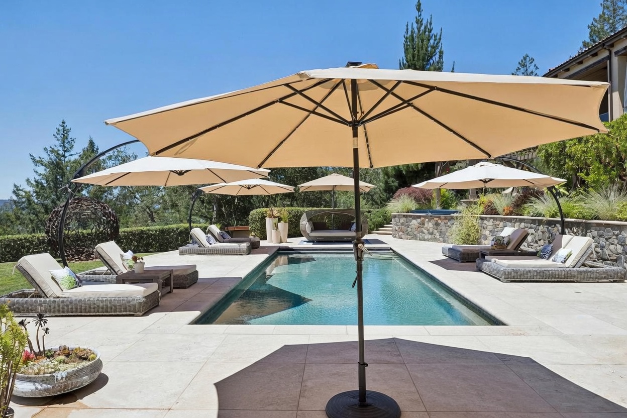 This expansive outdoor living space features a rectangular swimming pool surrounded by a stone patio and multiple lounge areas. Several large umbrellas provide shade for the dining set in the foreground and the various chaise lounges positioned around the water. The setting is framed by manicured hedges and mature trees, creating a private and serene atmosphere perfect for entertaining.