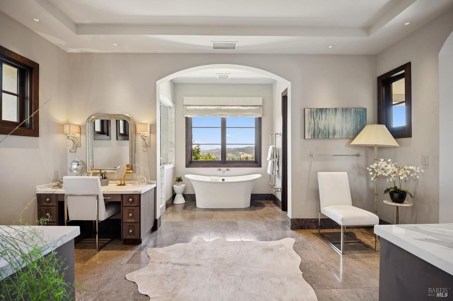 This luxurious primary bathroom features a central freestanding soaking tub positioned under an arched alcove with a scenic window view. To the left, a vanity area with a mirror and chair provides a dedicated space for grooming, while a white chair and floor lamp sit to the right. The room is finished with neutral-toned walls, stone flooring, and a large, light-colored hide rug, creating an elegant and serene atmosphere.