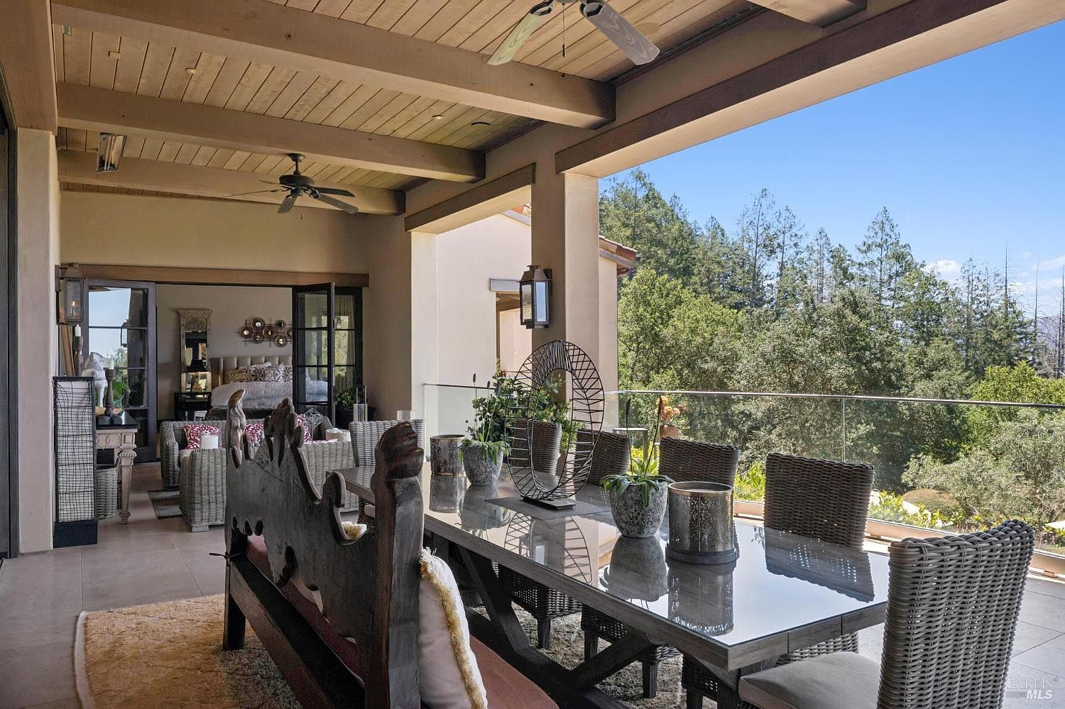 This expansive covered patio offers a seamless transition between indoor and outdoor living, featuring a large dining table and a rustic wooden bench. The space is framed by a glass railing that provides unobstructed views of the surrounding lush forest canopy. With its wood-paneled ceiling, ceiling fans, and integrated lighting, this area is designed for comfortable al fresco dining and relaxation.