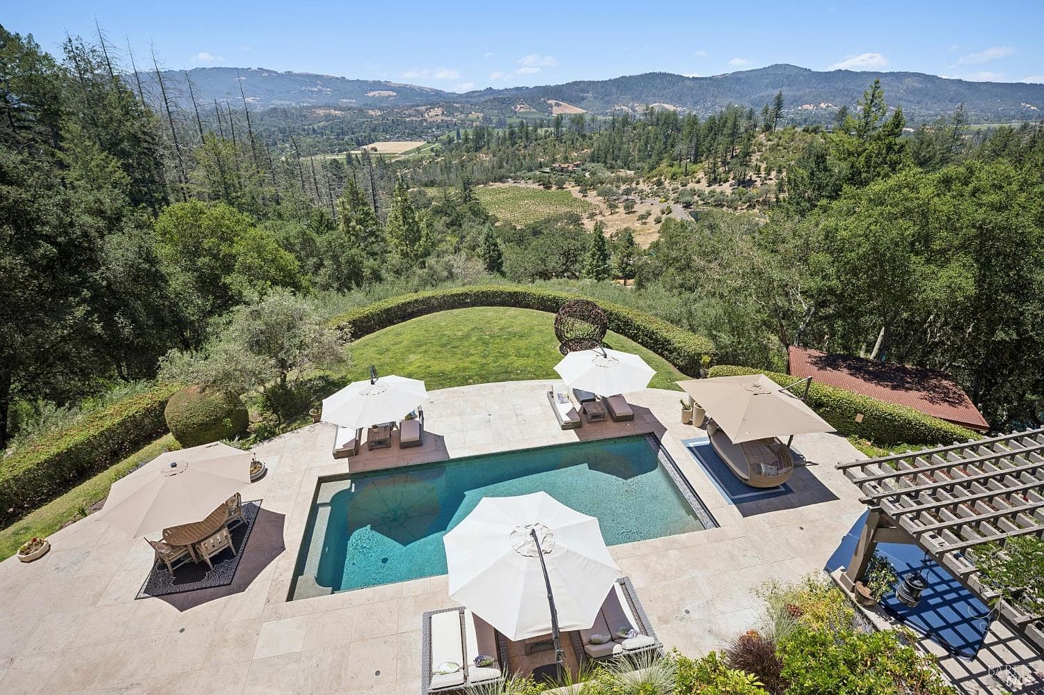 This high-angle aerial view captures a luxurious outdoor living space centered around a rectangular swimming pool set into a stone-paved terrace. The area is furnished with multiple lounge chairs, umbrellas, and a shaded pergola, all overlooking a manicured lawn and a sprawling, scenic valley landscape. The setting offers a serene and sophisticated atmosphere, perfect for outdoor entertaining with panoramic views of the surrounding hills and trees.