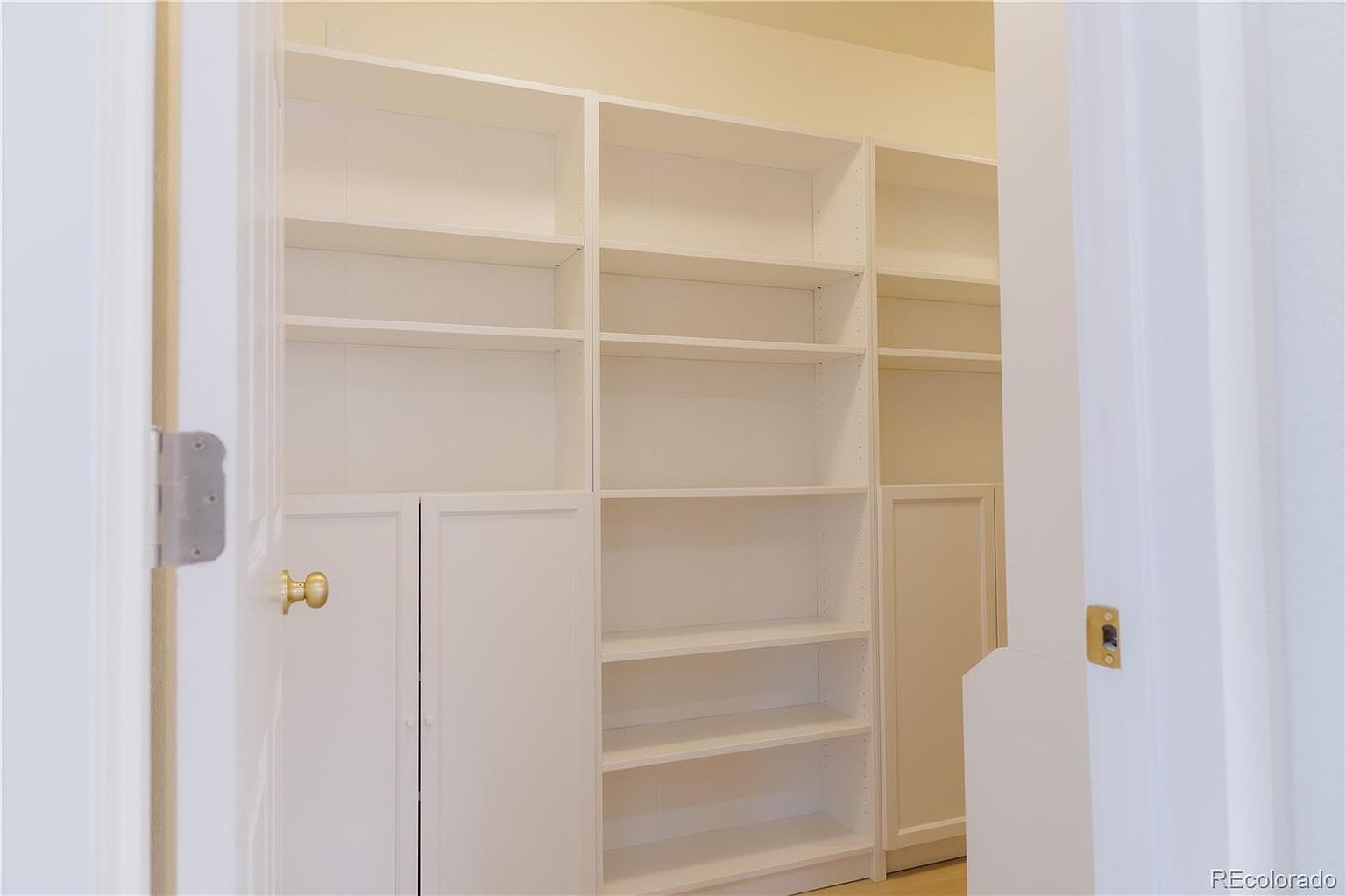 The image showcases a spacious walk-in closet with custom built-in shelving and cabinetry, all finished in a clean white color. The closet is well-organized and appears to offer ample storage space. The view is framed by a partially open door on either side, adding depth and perspective.