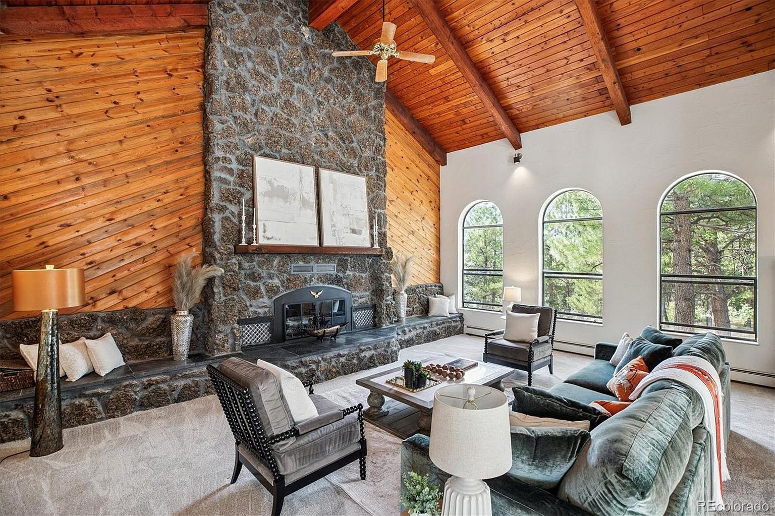 This is an interior shot of a living room featuring a stone fireplace, wood-paneled walls, and a vaulted ceiling with exposed beams. The room is furnished with a plush green sofa, armchairs, and a coffee table, creating a cozy and inviting atmosphere. Large arched windows provide natural light and views of the surrounding landscape.